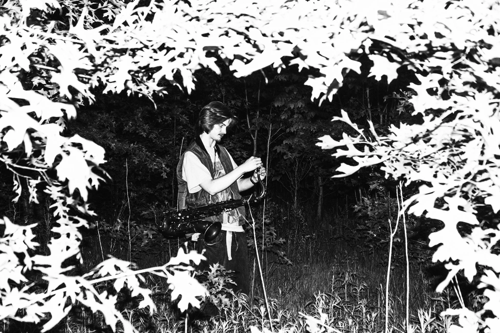 A person standing outdoors among dense trees and leaves, holding a saxophone.