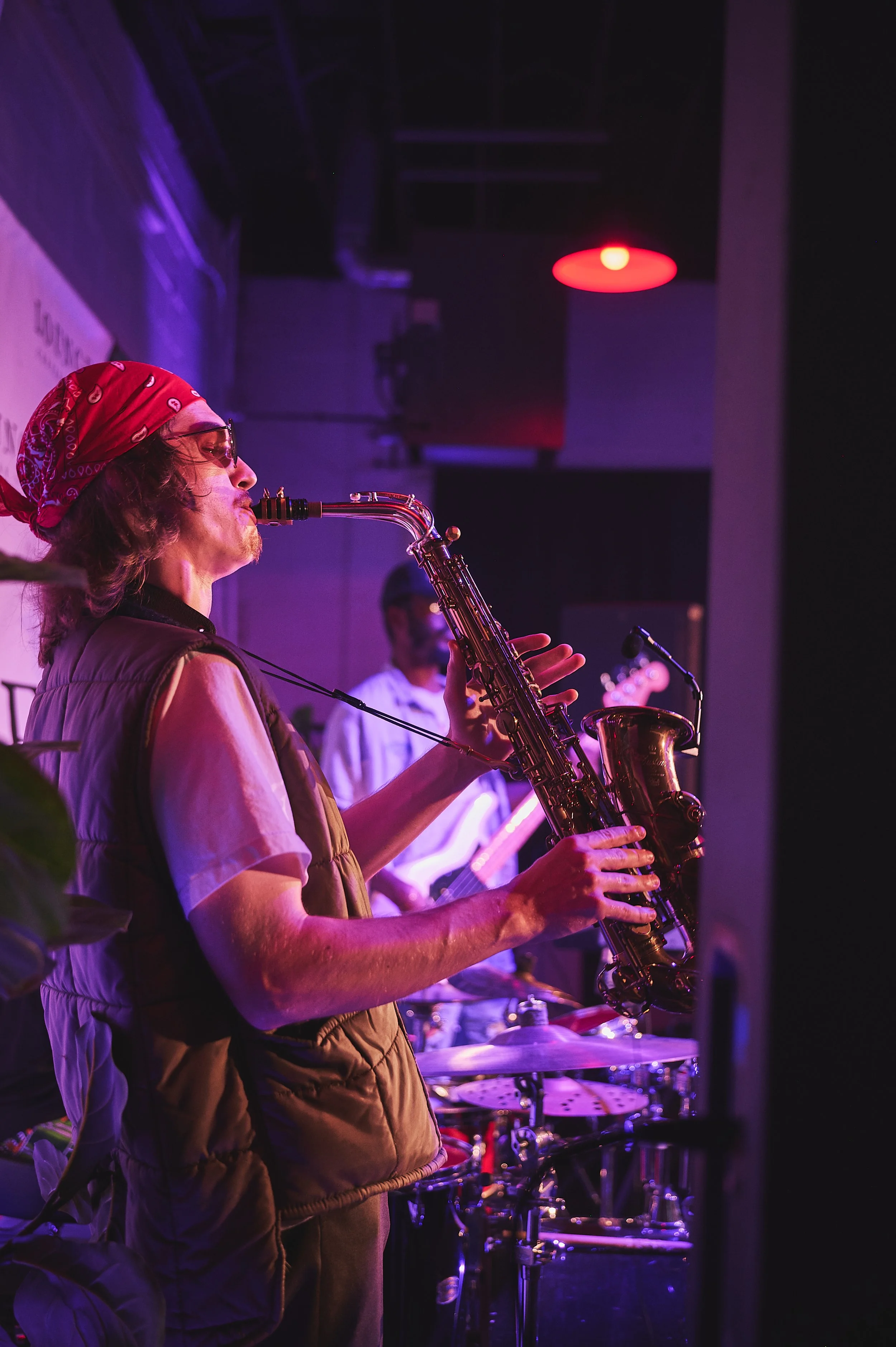 A musician with long hair, wearing a red bandana, glasses, and a sleeveless vest, playing a saxophone on stage under purple and red lighting.