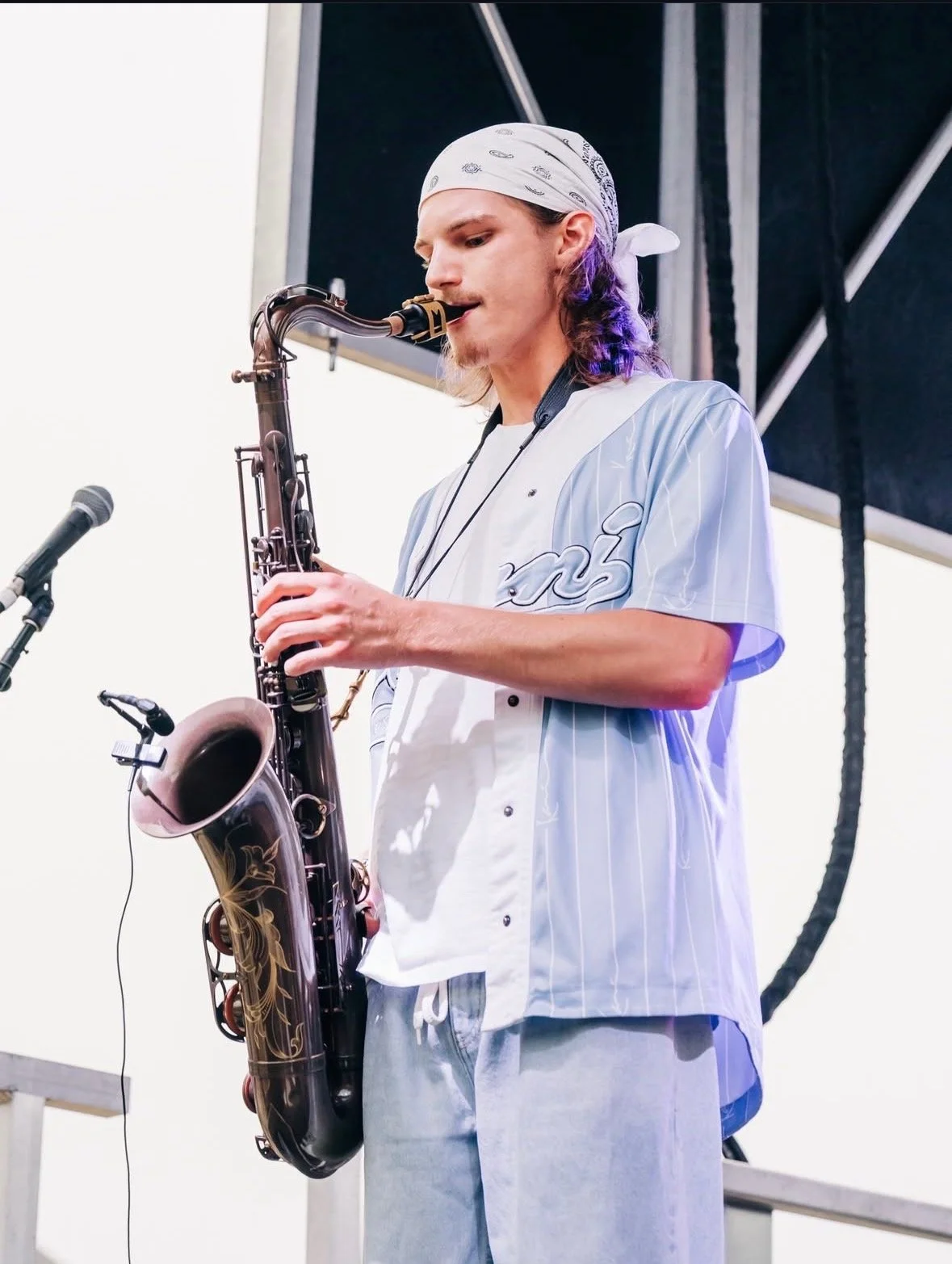 Young man wearing a white bandana and a light blue and white striped shirt playing a saxophone on stage.