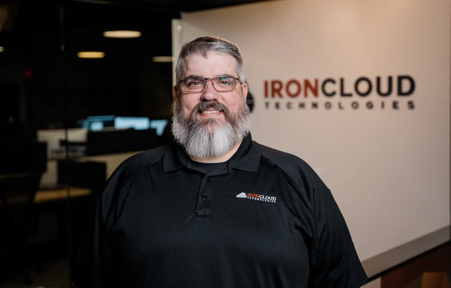 A man with glasses and a gray beard standing in front of a wall with the IronCloud Technologies logo in an office environment.