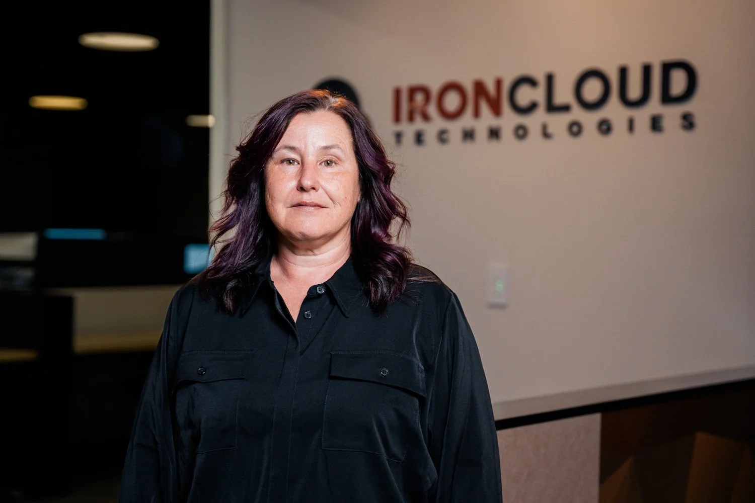 A woman with shoulder-length dark purple hair stands in front of a wall with a logo that reads 'IRONCLOUD TECHNOLOGIES' in a modern office setting.