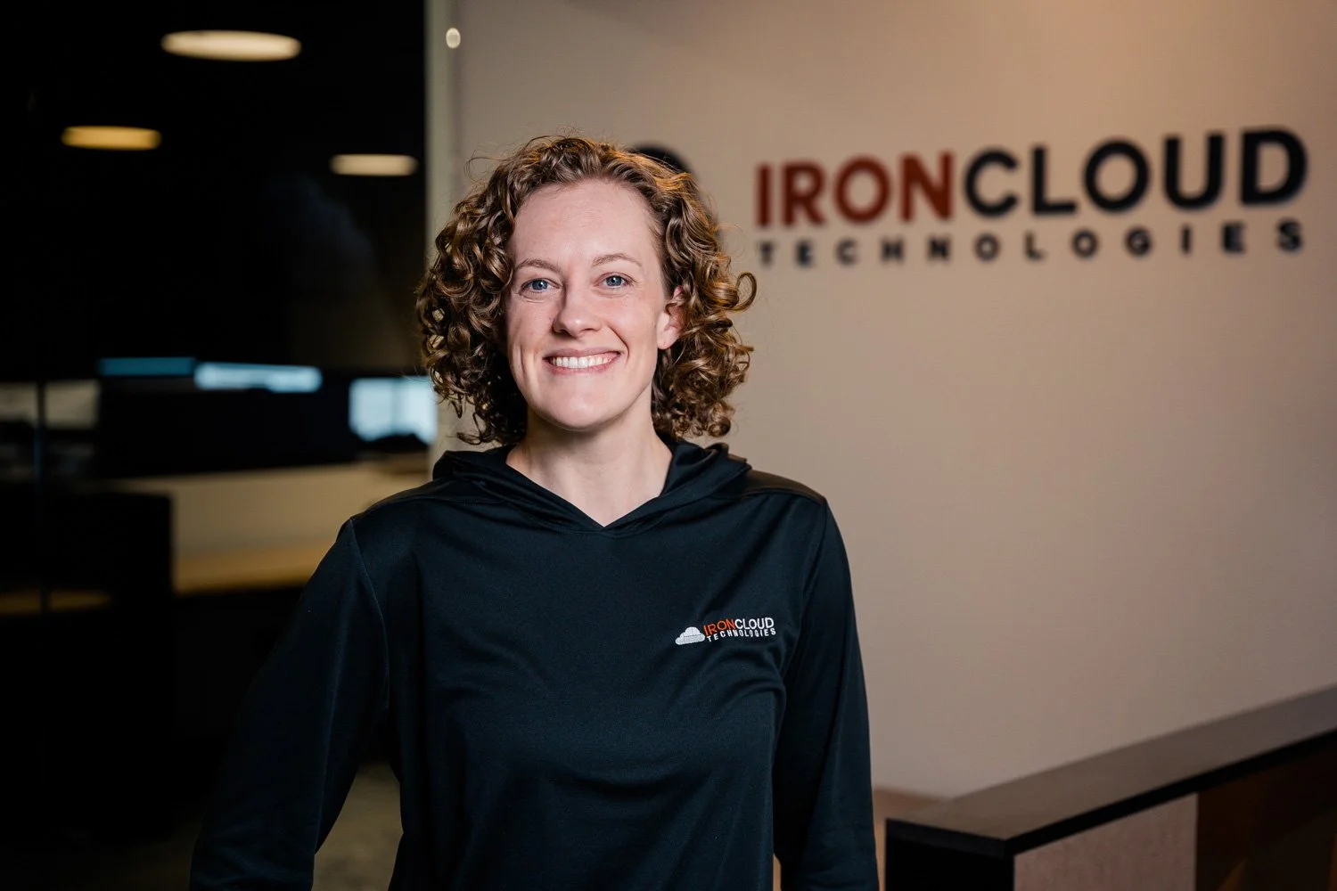 A woman with curly hair smiling, wearing a black hoodie with a logo that says 'IronCloud Technologies,' standing in an office with a blurred background.