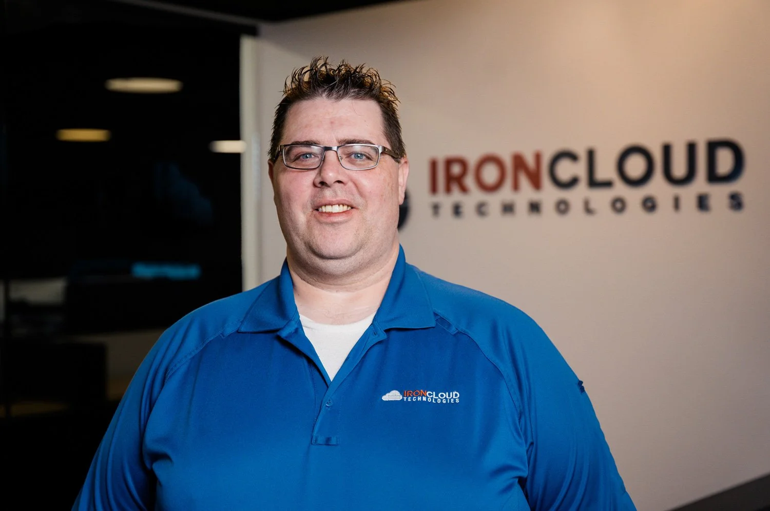 A man with glasses in a blue polo shirt stands indoors in front of a wall with 'IronCloud Technologies' logo. The logo is somewhat blurred but visible.
