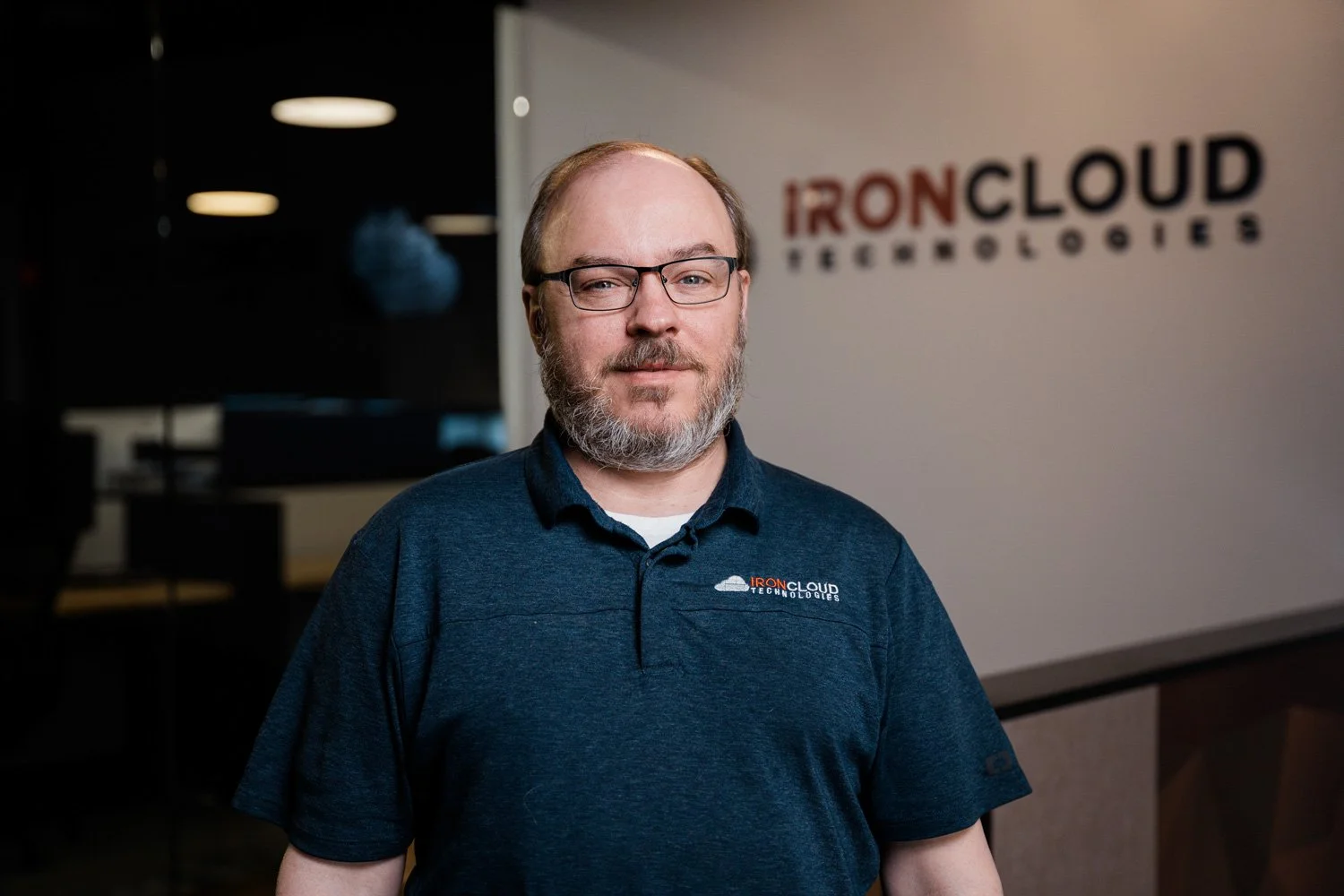 Man with glasses and a beard wearing a navy blue polo shirt with a company logo standing in front of a wall with the IRONCLOUD Technologies logo.