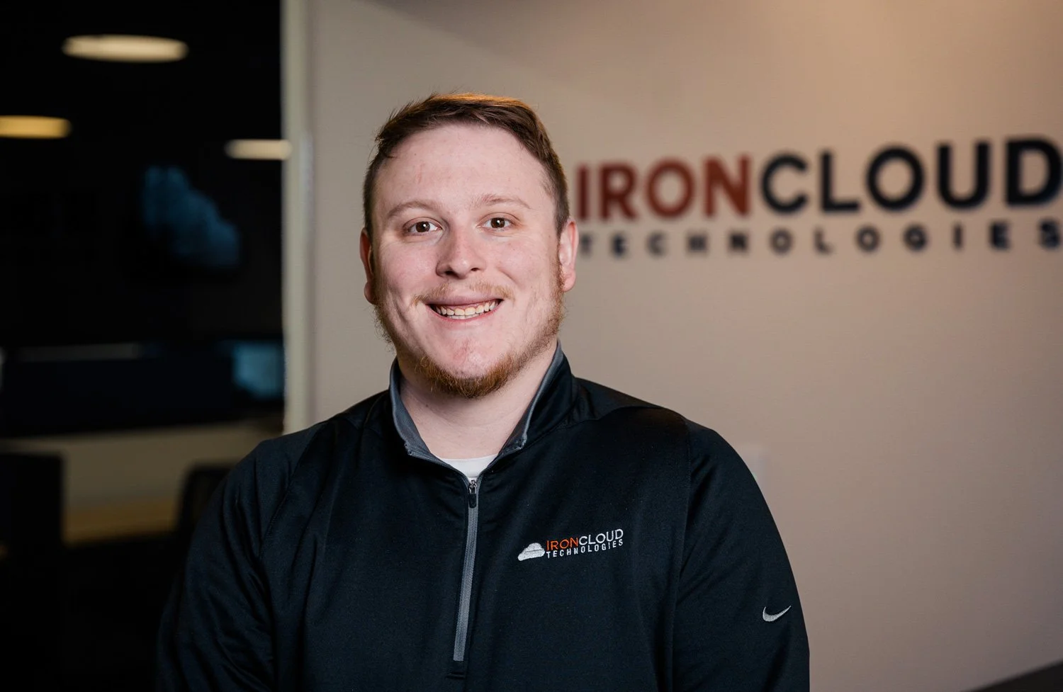 Young man with short brown hair and beard smiling in front of a wall with 'IRONCLOUD TECHNOLOGIES' logo, wearing a black 'IronCloud Technologies' jacket.