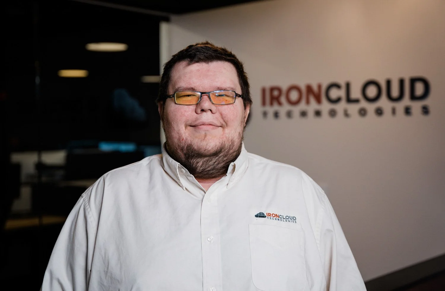A man with glasses, short dark hair, and a beard smiling, wearing a white collared shirt with IronCloud Technologies logo, standing in front of a wall with a blurred IronCloud Technologies sign.