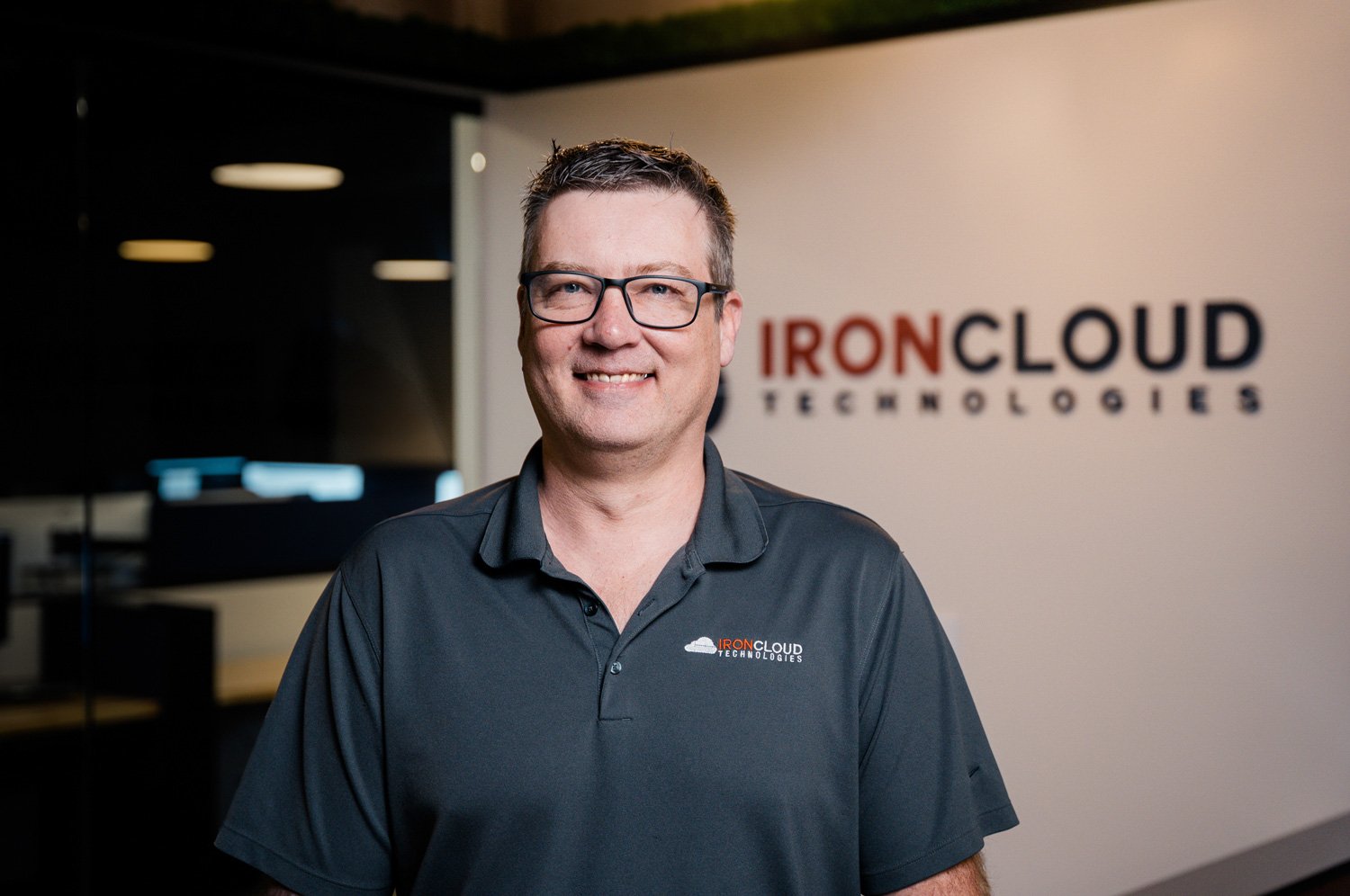A man wearing glasses and a black polo shirt with an IronCloud Technologies logo stands in front of a wall with the same logo, smiling at the camera in an office or tech environment.