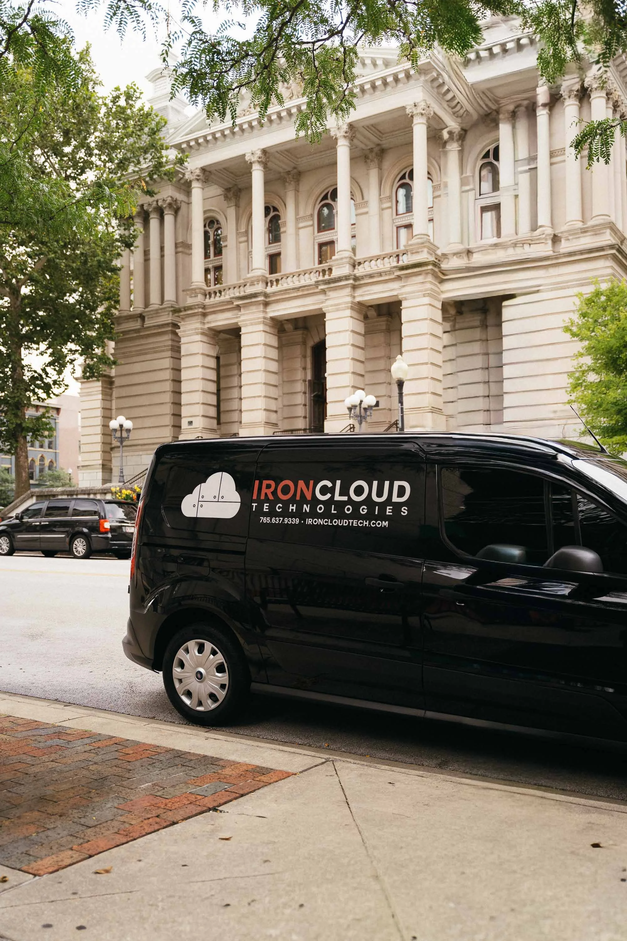 A black van with the logo and name of IronCloud Technologies parked on a city street in front of a historic building with columns and ornate architecture. There are other cars parked nearby, and trees line the street.