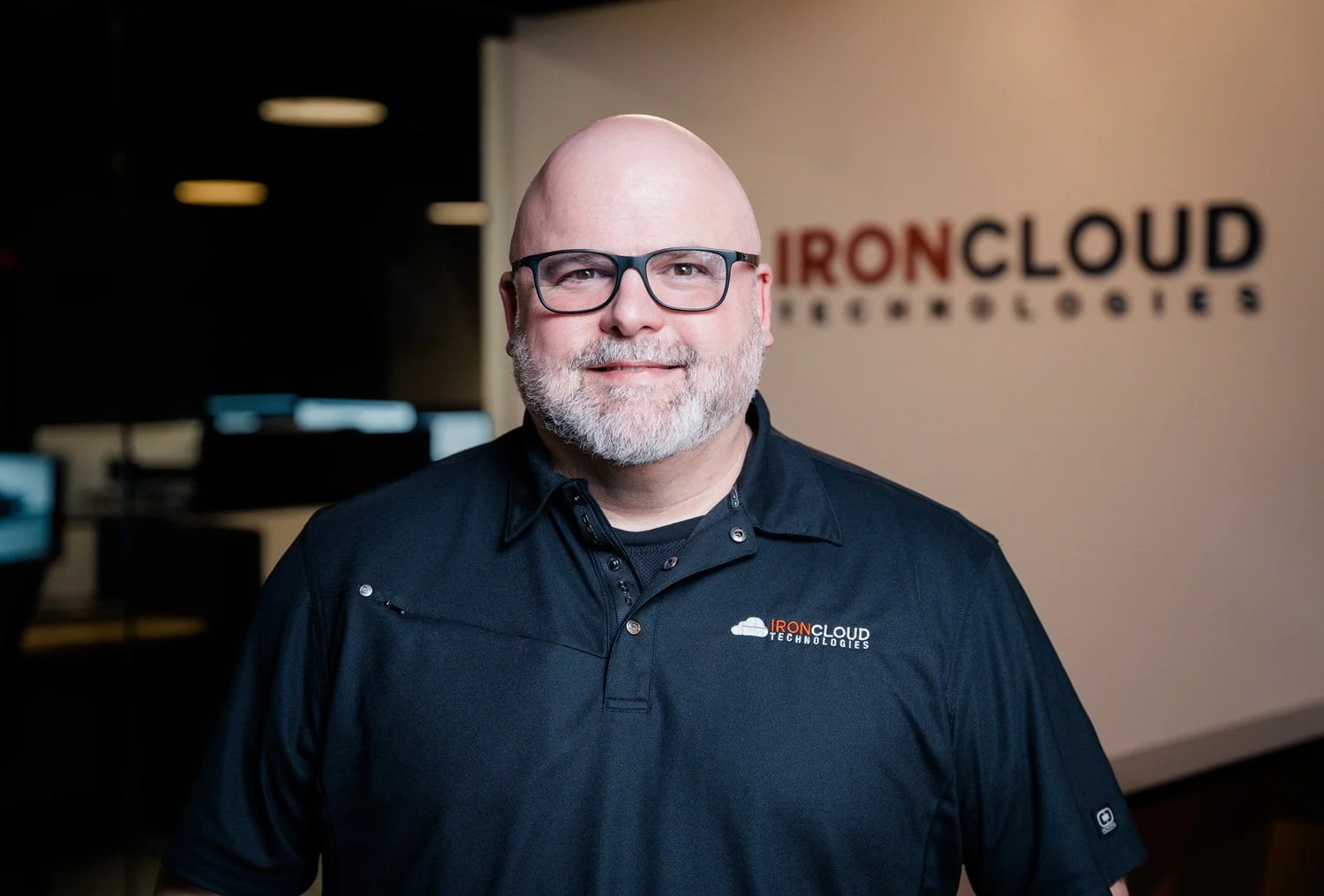 Man with glasses and a beard smiling, wearing a black polo shirt with the IronCloud Technologies logo, standing indoors with the company's sign visible in the background.