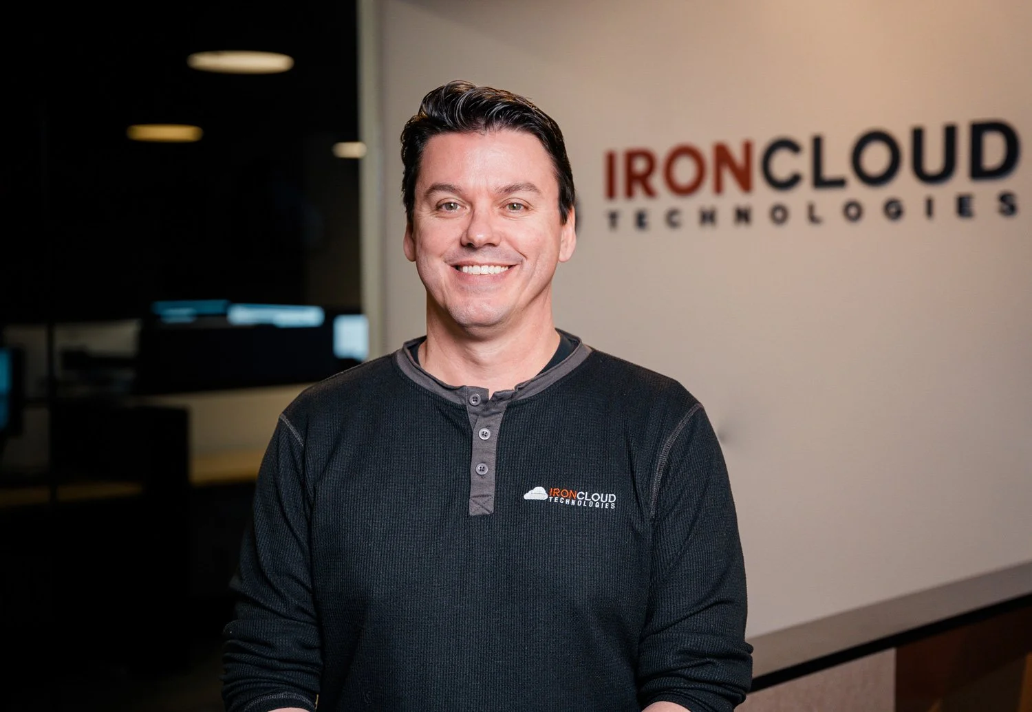 A man smiling in front of a wall with the Ironcloud Technologies logo, wearing a black sweatshirt with the same logo.