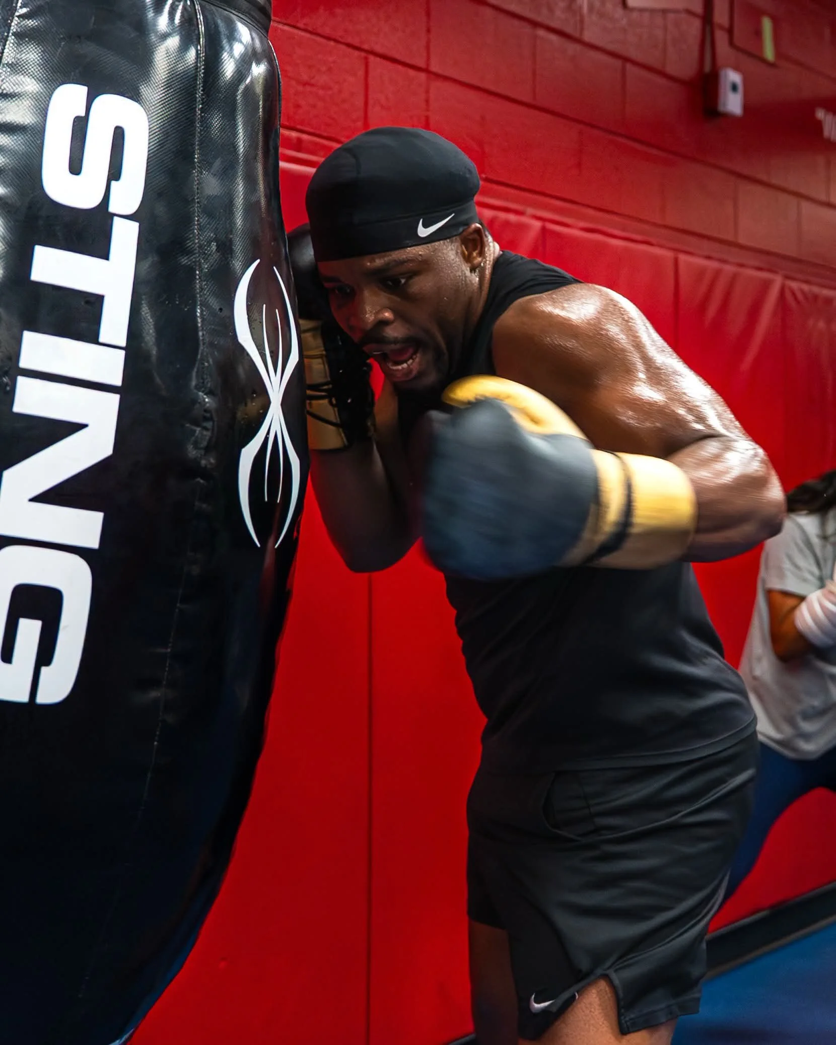 A man with a black Nike headband and black athletic clothing is practicing boxing in a gym, leaning against a red padded wall and hitting a black punching bag with white text.