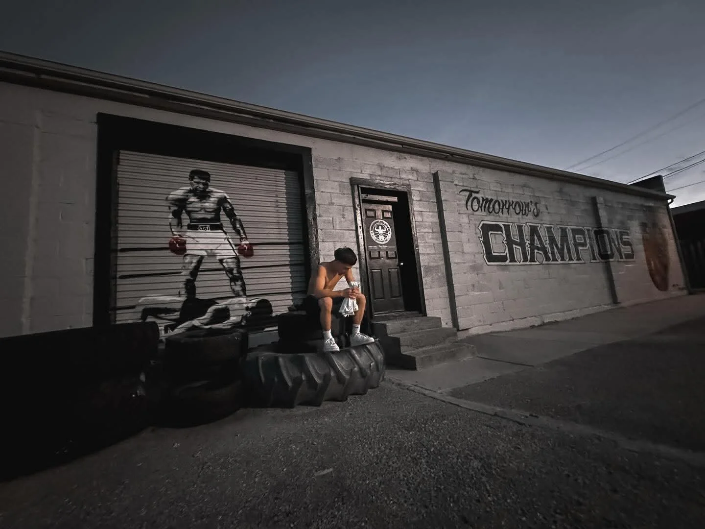 A young man is sitting on tires outside a building with a mural of a boxer, Mohammad Ali, painted on peeling metal garage doors. The wall has the words 'Tomorrow's Champions' painted on it.