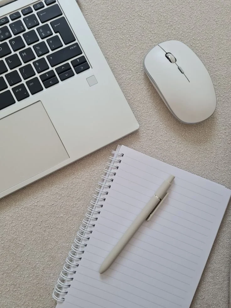 A laptop, a wireless mouse, a notepad, and a pen on a beige surface.