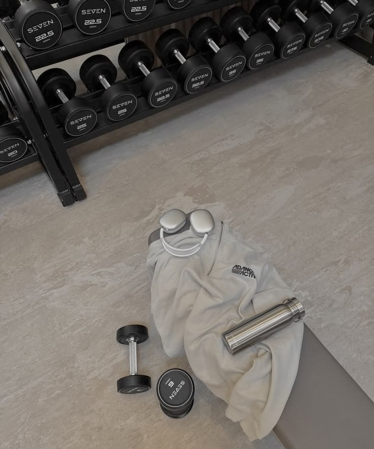 Workout gear including dumbbells, a water bottle, wireless earbuds, a towel, and a small container on the floor near a rack of dumbbells.