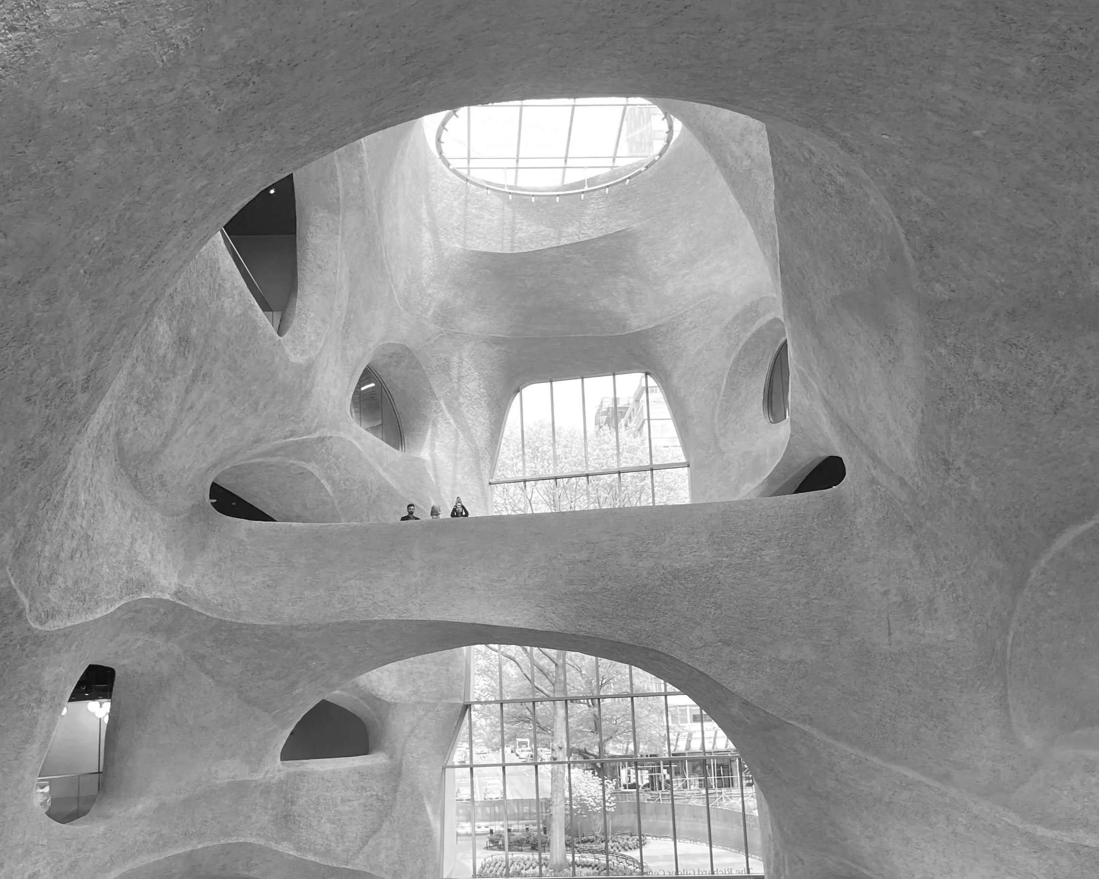 Interior view of a modern architectural building with concrete walls, large windows, and an oval skylight at the ceiling, with three people standing near the bottom.