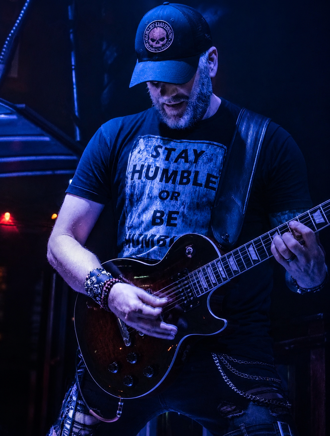 A man wearing a black cap with a skull logo, a black T-shirt with the phrase 'STAY HUMBLE OR BE HUMBLE' playing an electric guitar in a dark setting with blue lighting.