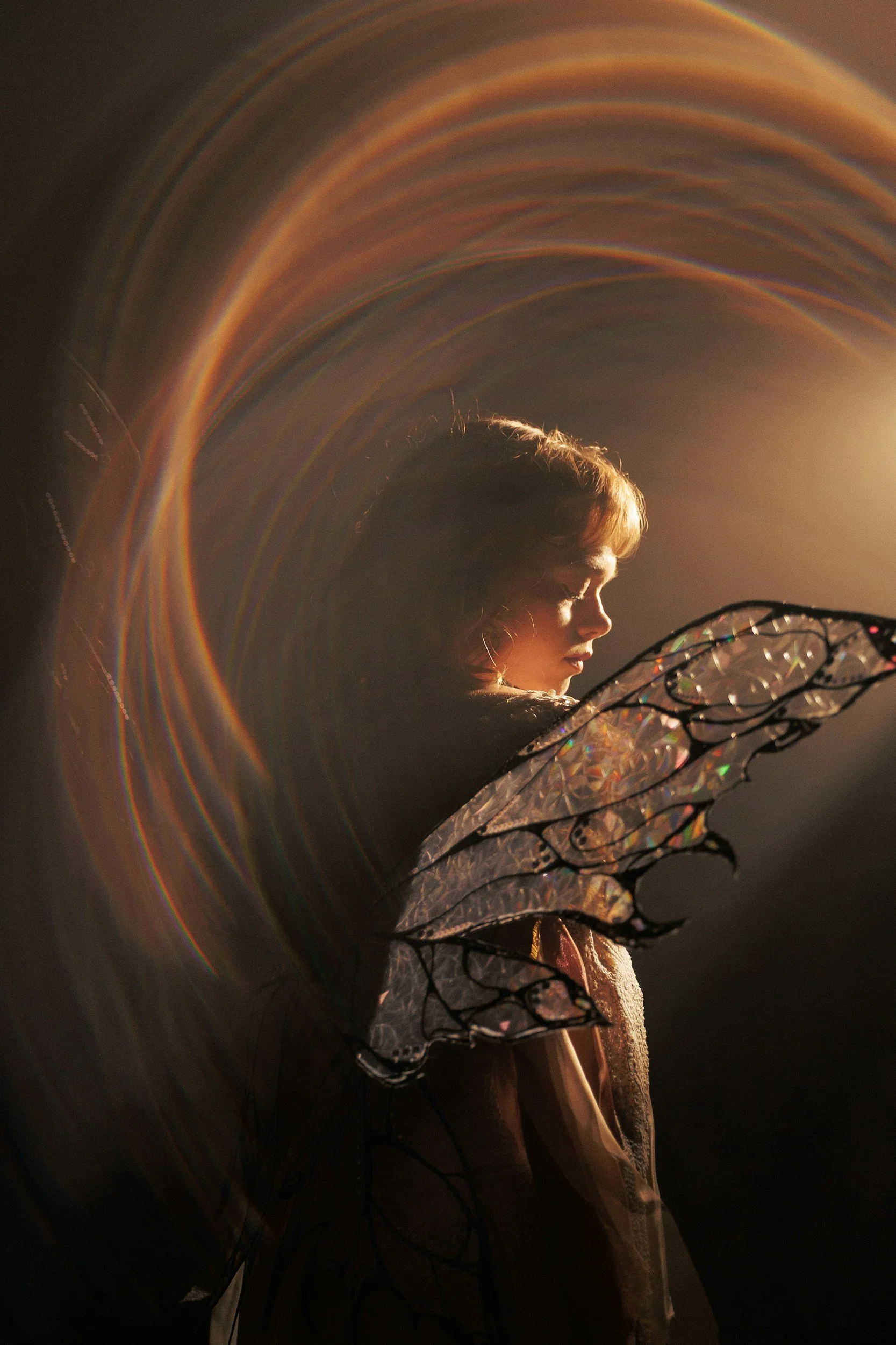 A girl with fairy wings lit by a warm light, with colorful light trails creating a rainbow arc around her head.