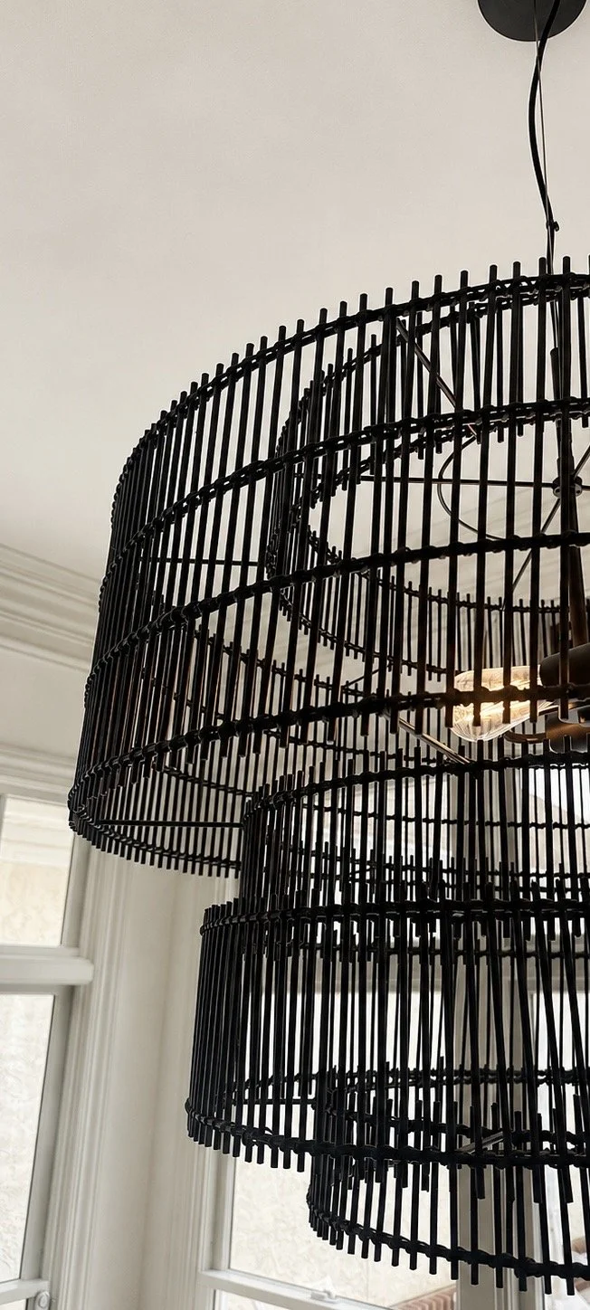 Close-up of a modern black metal chandelier with a tiered, open cylindrical design hanging in a room with white walls and windows.