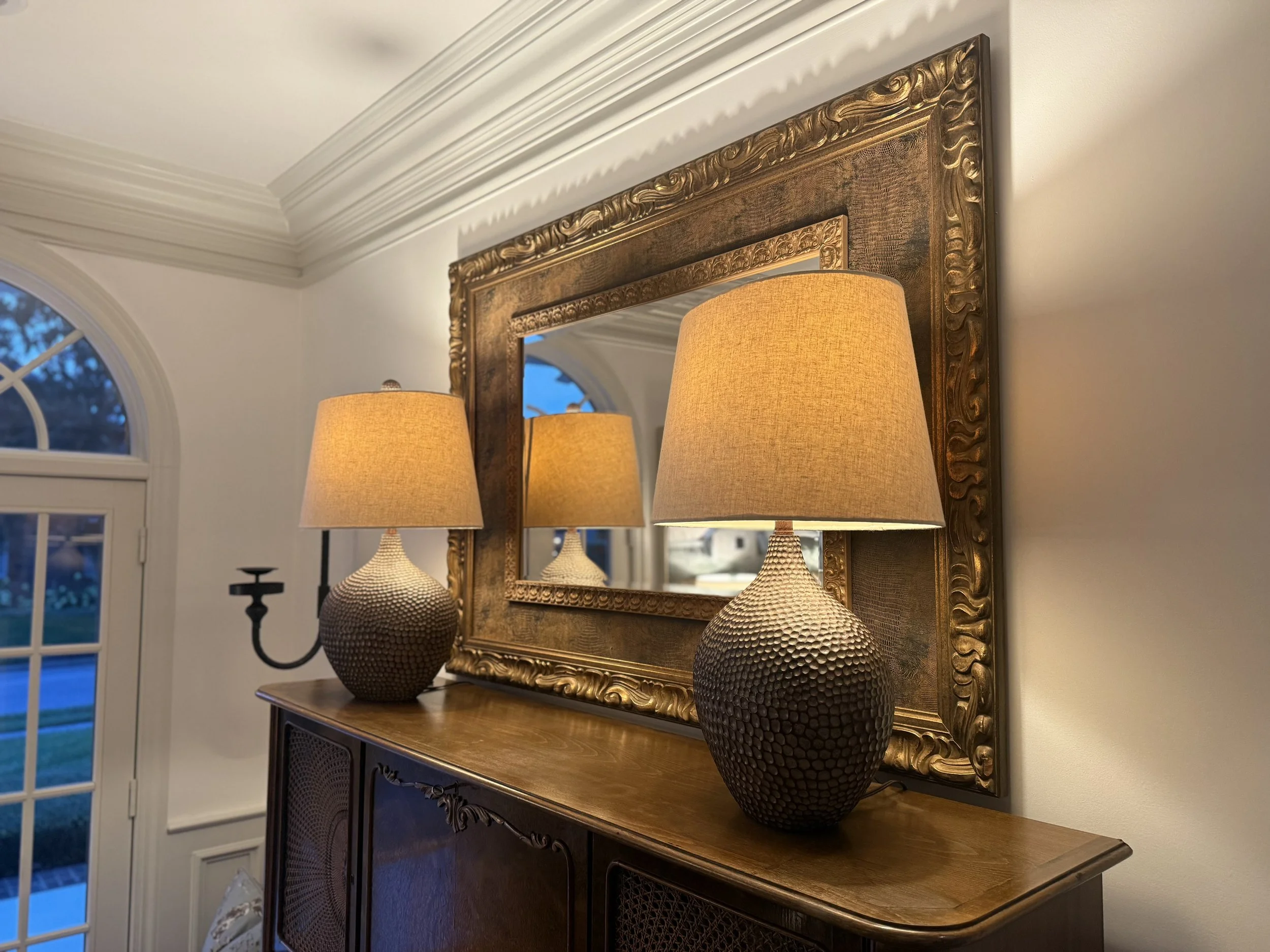 A decorative wooden sideboard with a mirror above it flanked by two ornate lamps with textured, dark brown bases and beige lampshades, inside a home with white walls and crown molding.