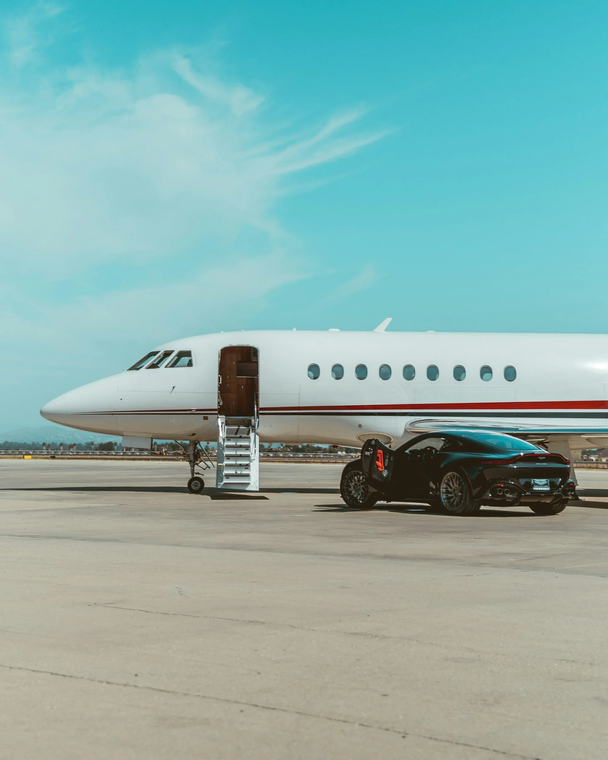 Part of a white private jet with a door open and metal stairs leading up to it, on the tarmac near a black sports car, under a partly cloudy sky.