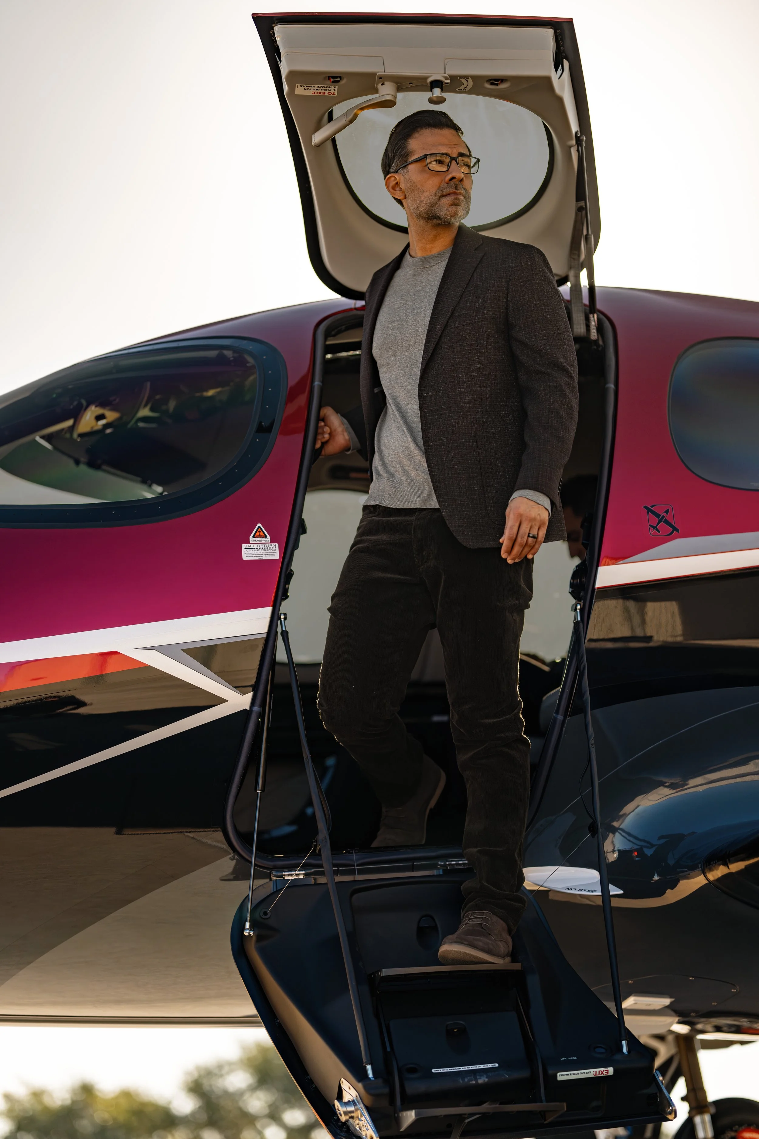 A man stands next to a vision jet, holding the open door while looking to his left. He is wearing glasses, a gray shirt, a dark blazer, and dark pants. The helicopter is black and some parts are gray and red.