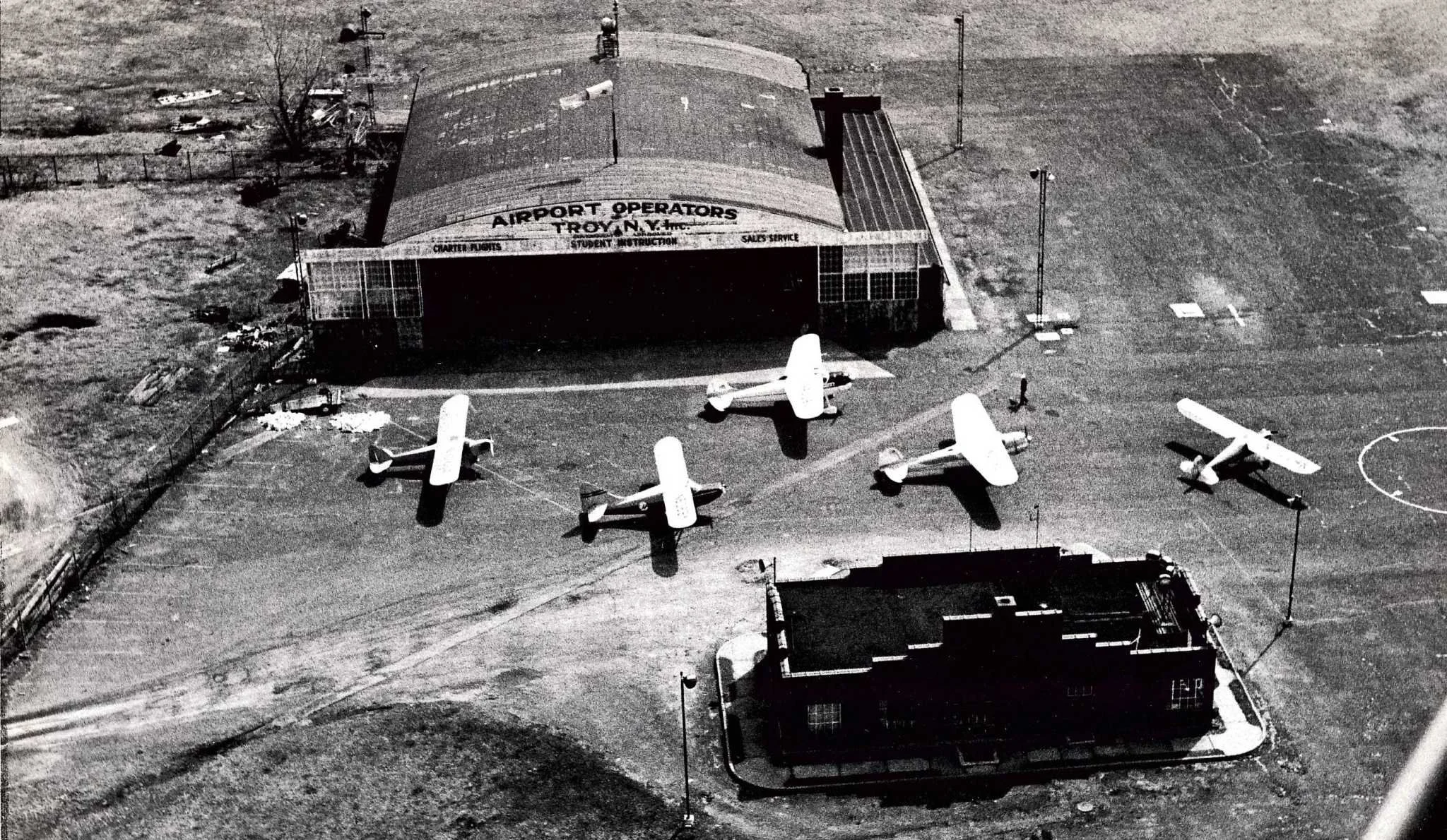 Black-and-white aerial photo of a small airport with four small aircraft parked on the tarmac. The terminal building has a sign reading "Airport Operators Troy N.Y." and offers charter flights, student instruction, and sales service.