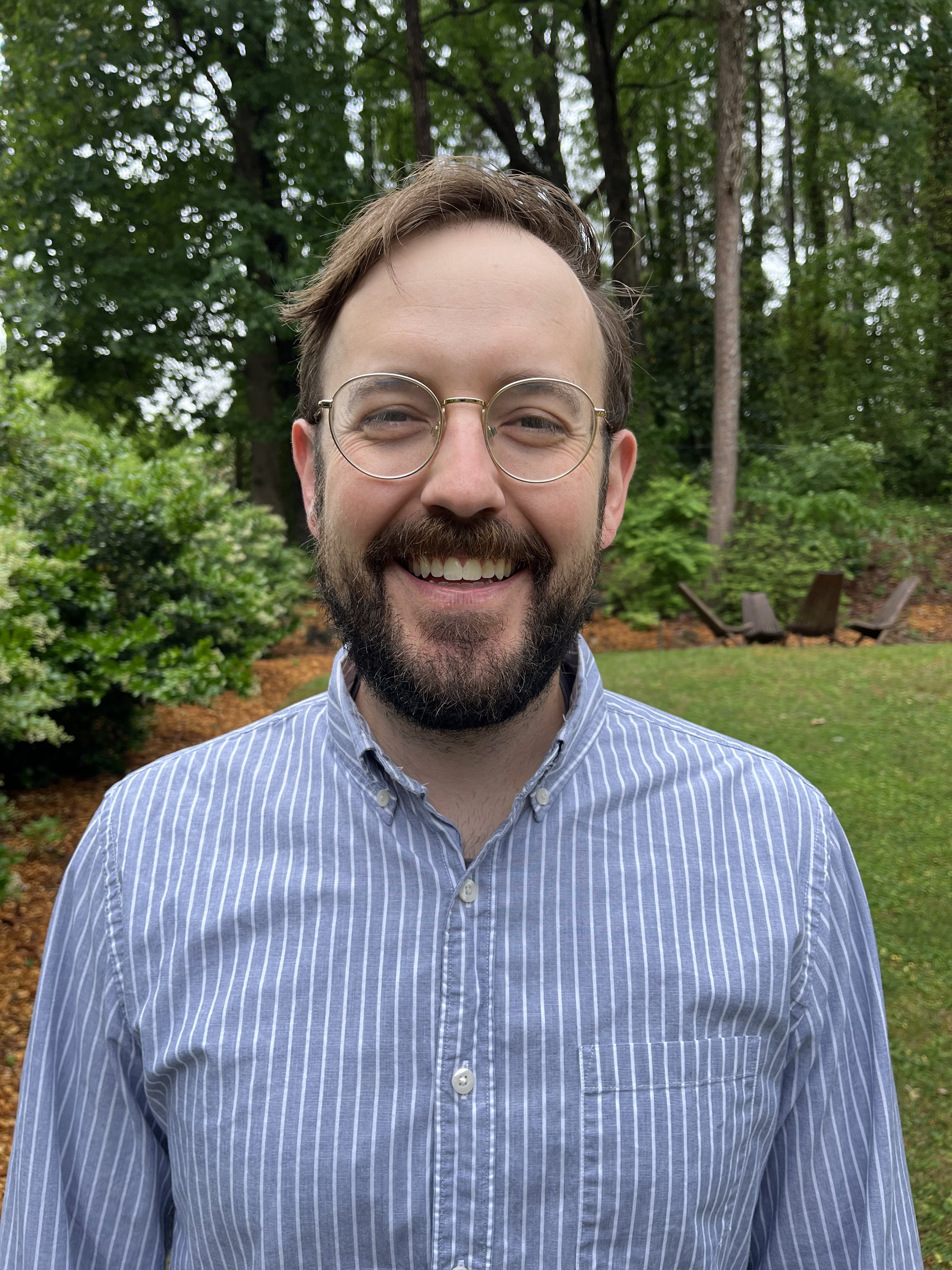 A smiling man with glasses and a beard, wearing a blue and white striped shirt, stands outside in a garden with green trees and bushes in the background.