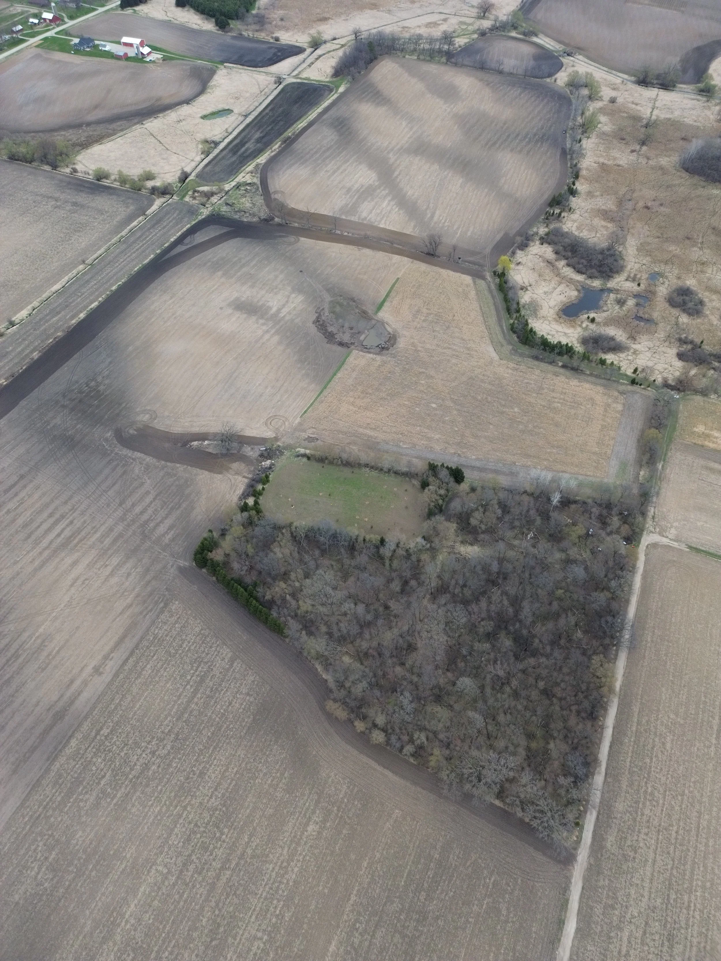 Aerial view of farmland with various plowed fields, some with crops, and patches of trees, roads, and small ponds.