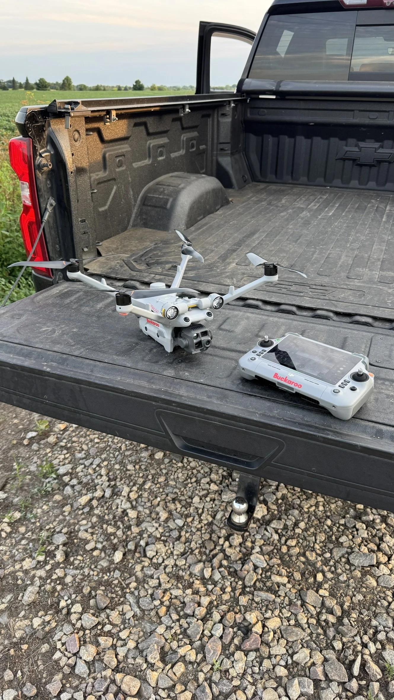 A white drone with four propellers and a camera is on the tailgate of a black pickup truck. Alongside the drone, there is a white remote control with the brand name Buckaroo. The truck's bed is empty and the background shows green fields and a cloudy sky.