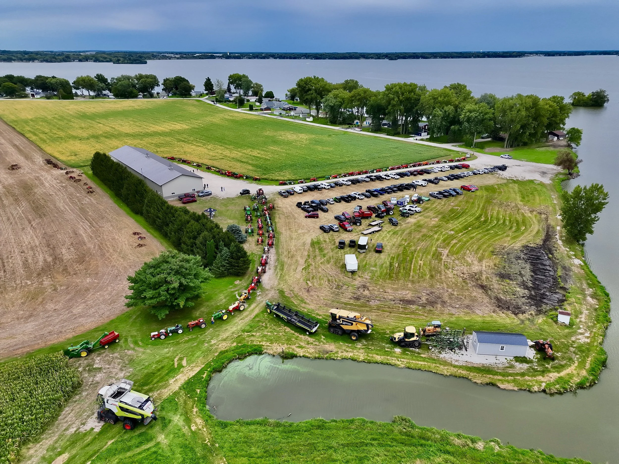 Aerial view of a rural area with a mix of farmland, a small pond, and a residential neighborhood near a large body of water, featuring parked cars, tractors, and farm equipment.
