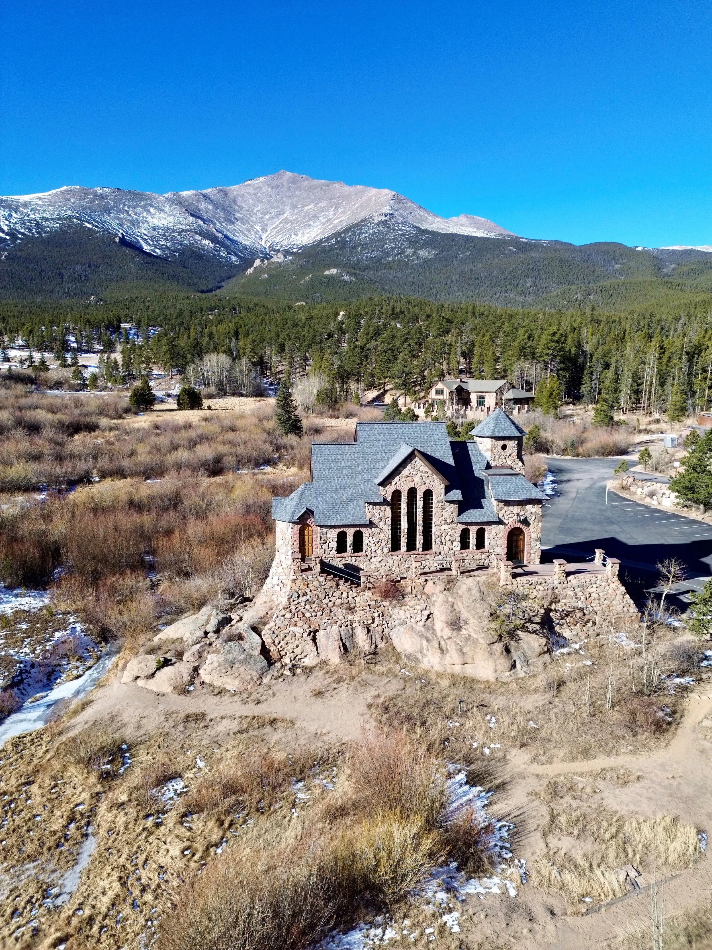Scenic view of a mountainous landscape with a stone chapel, pine trees, a clear blue sky, and snow patches on the ground.