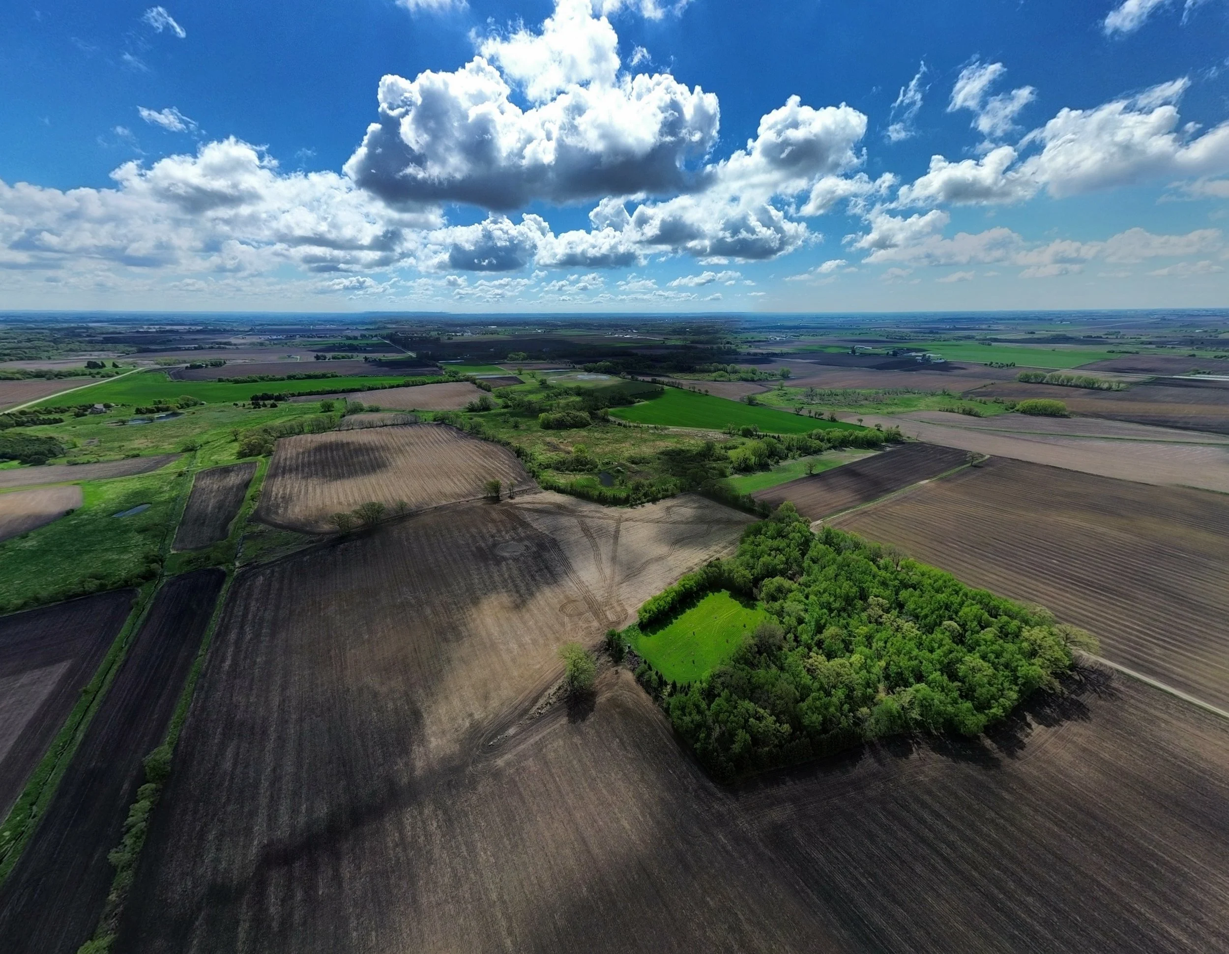 Aerial view of a rural landscape with patchwork fields, green pastures, and scattered trees under a partly cloudy sky.