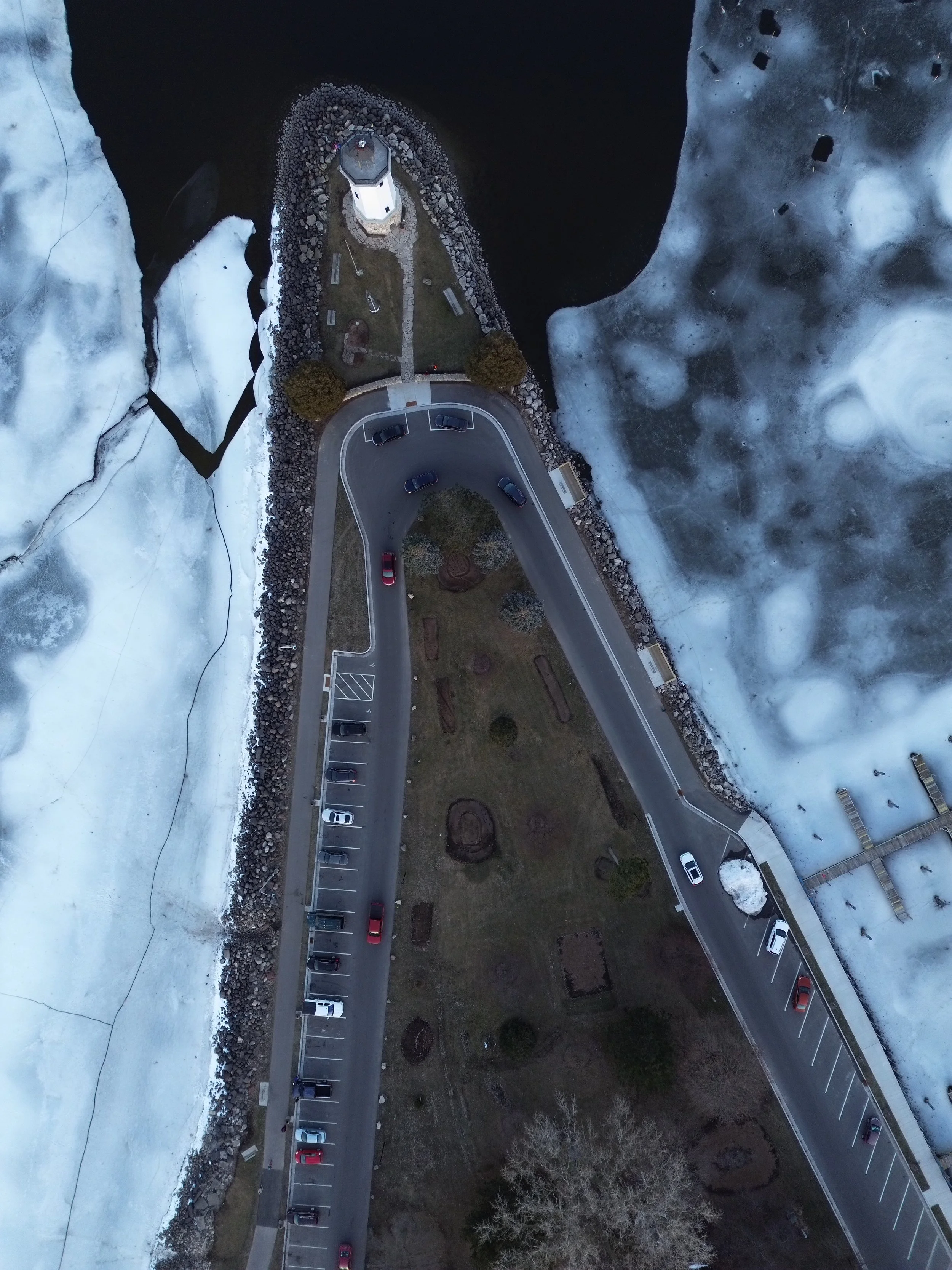 An aerial view of a lighthouse situated on a small peninsula surrounded by icy water, with parking area and roads leading to it, and snow-covered landscape in the surroundings.