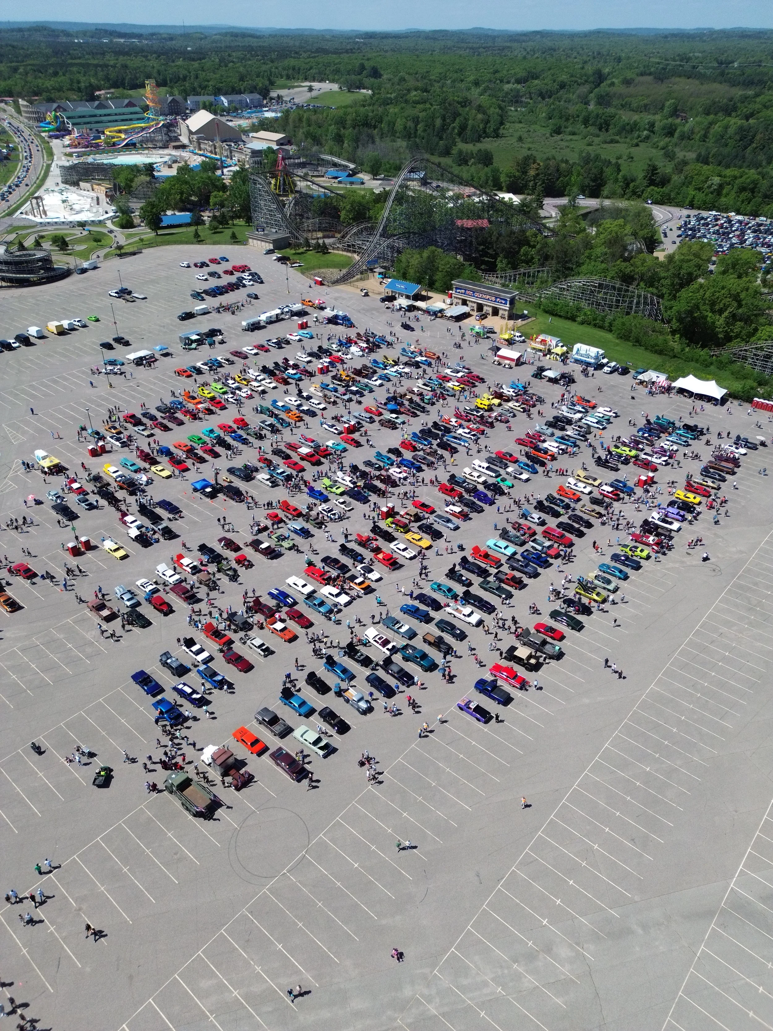 Aerial view of a parking lot filled with colorful cars, surrounded by trees and a theme park with roller coasters and rides in the background.