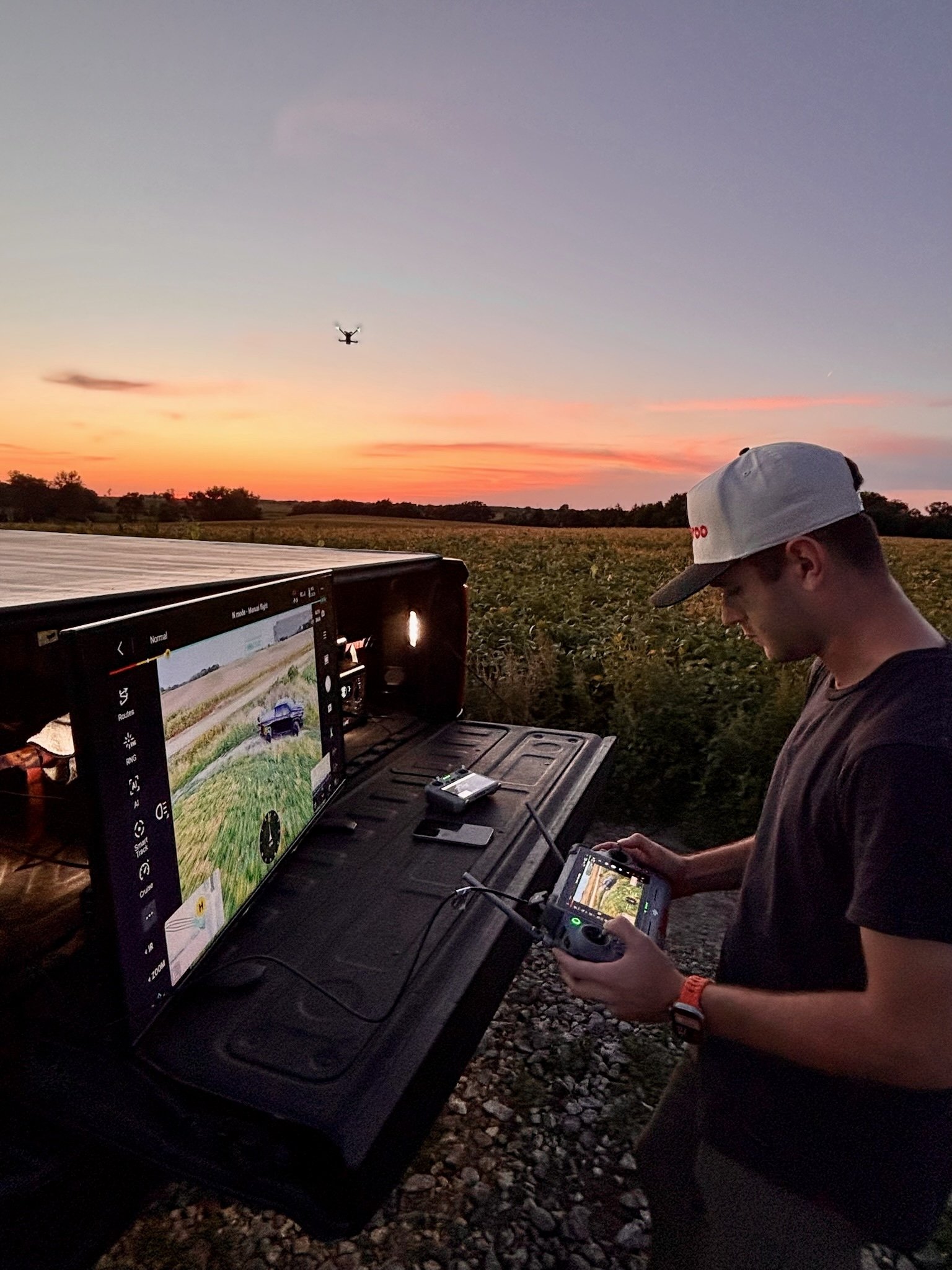 A person wearing a white cap and dark shirt is operating drone control equipment with a screen displaying a vehicle on a field, during sunset, with a drone flying in the sky.