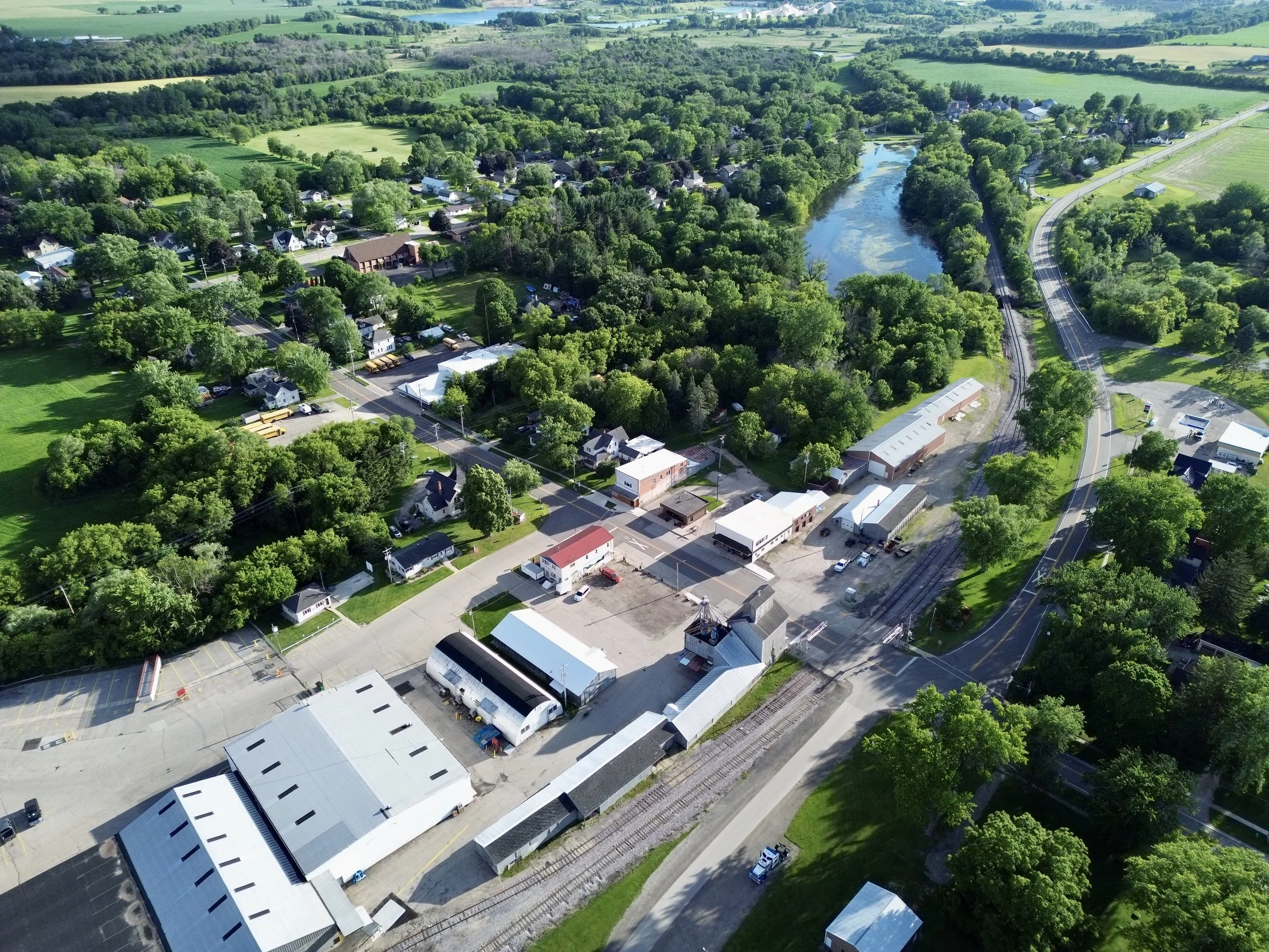 Aerial view of a small town with residential houses, commercial buildings, surrounded by lush green trees, fields, and a river with a train track running alongside it.