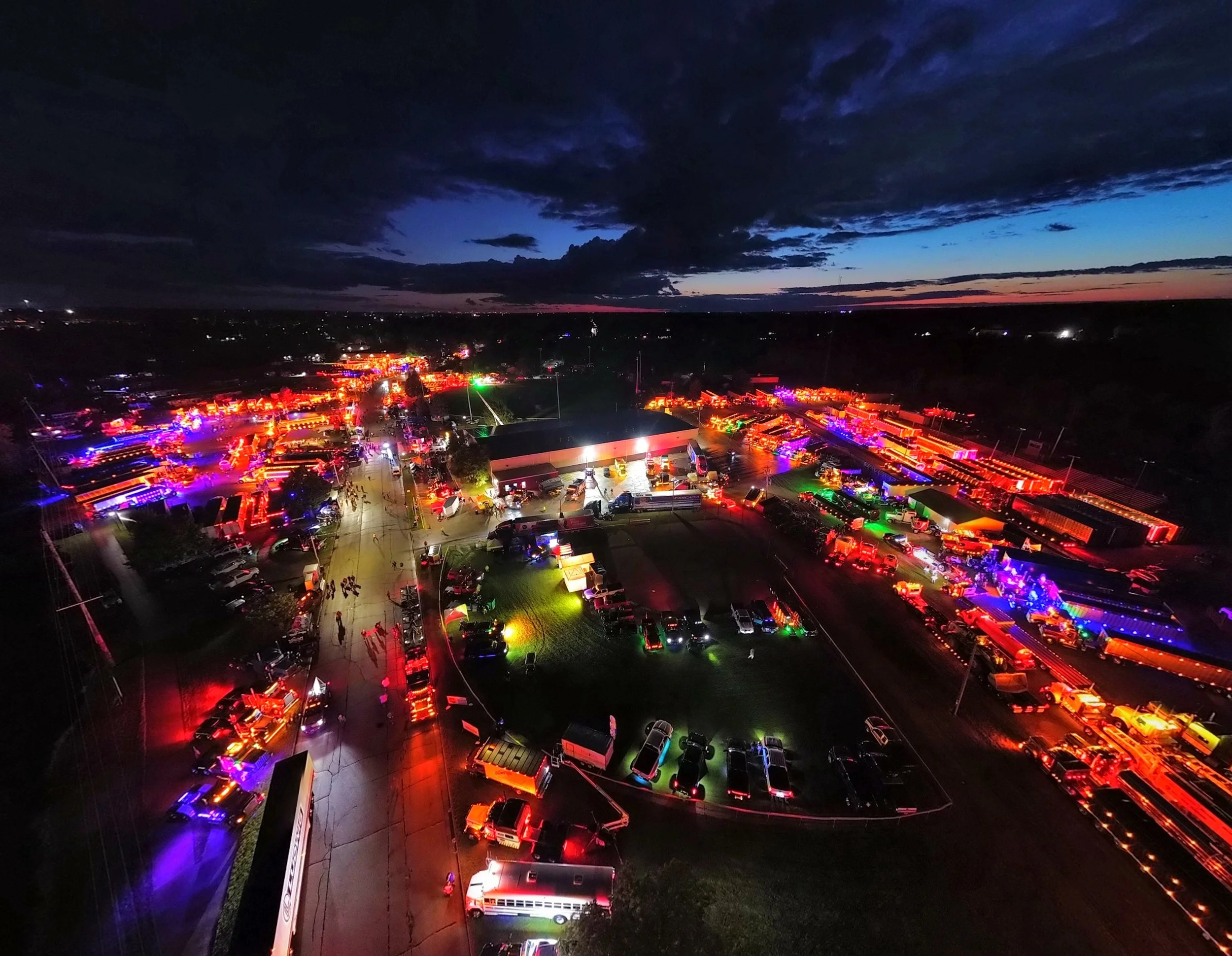 Aerial view of a nighttime fairground or carnival with colorful neon lights illuminating tents, rides, and booths, under a cloudy sky with a faint sunset in the background.