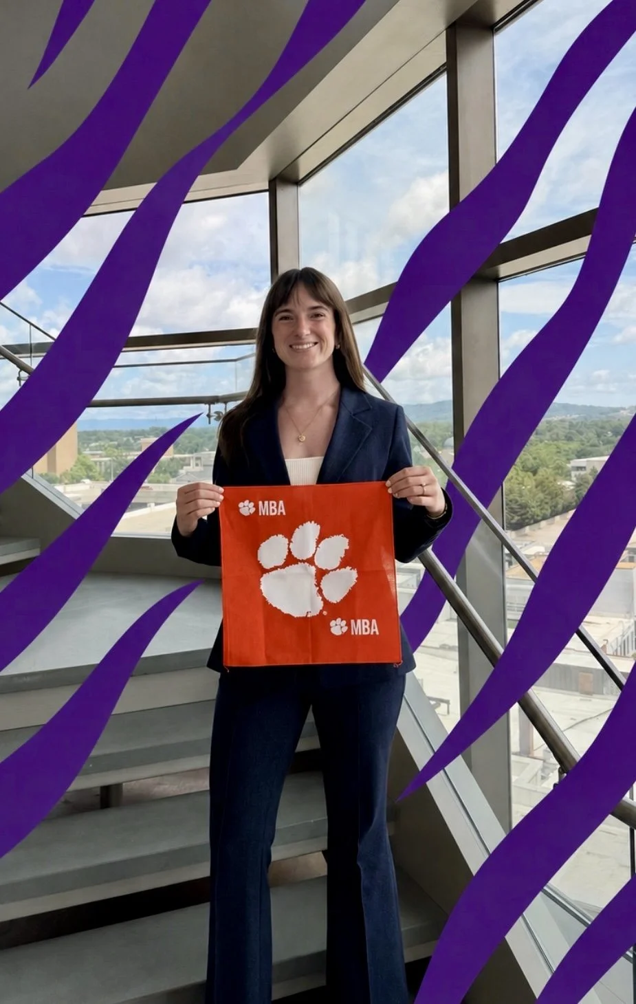 Woman in professional attire holding an orange Clemson University MBA bag inside a modern building with large glass windows and a scenic outdoor view.