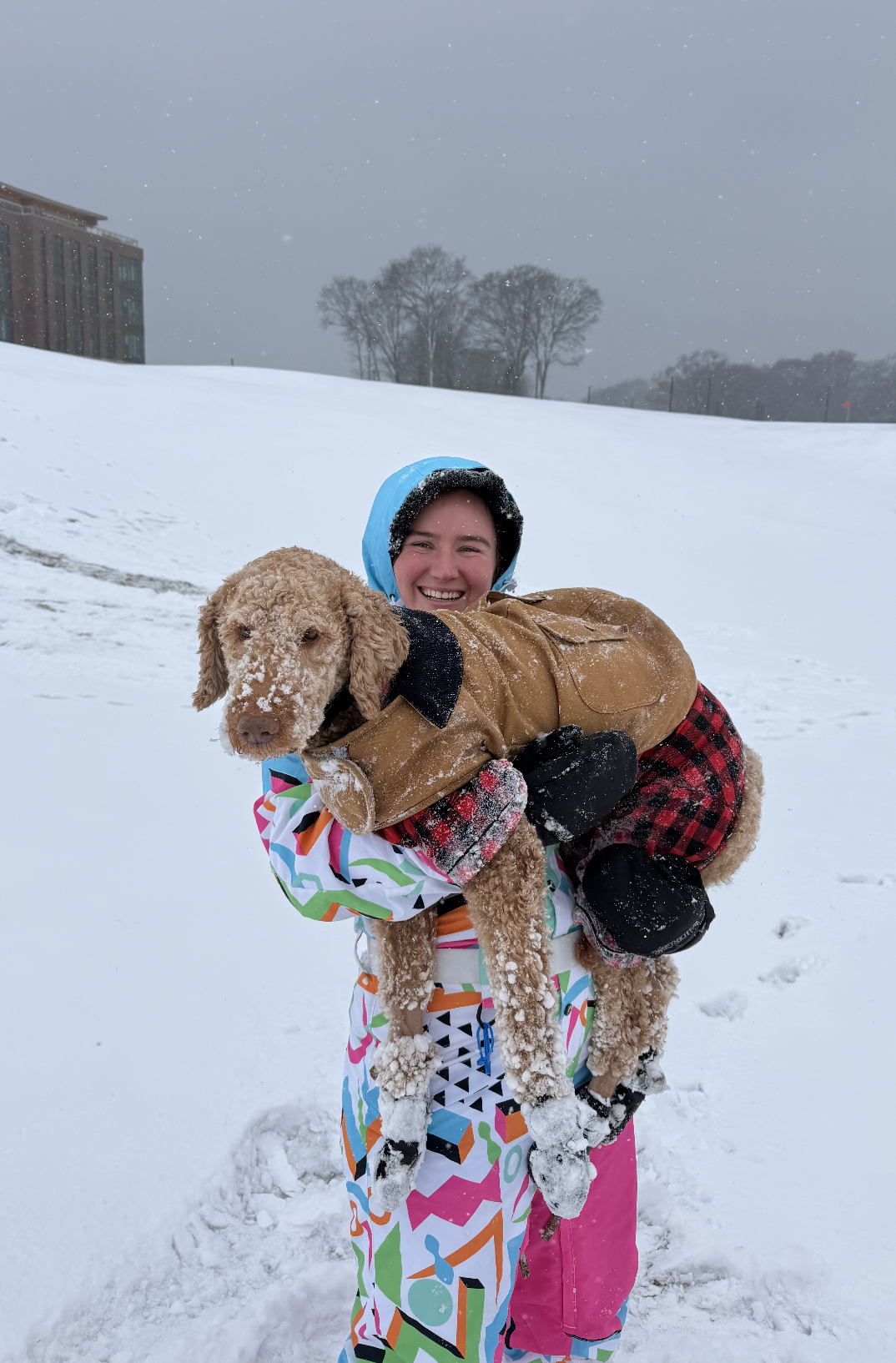 A woman in colorful winter clothing and a blue hood hugging a large, curly-haired dog wearing a brown coat in a snowy outdoor setting.