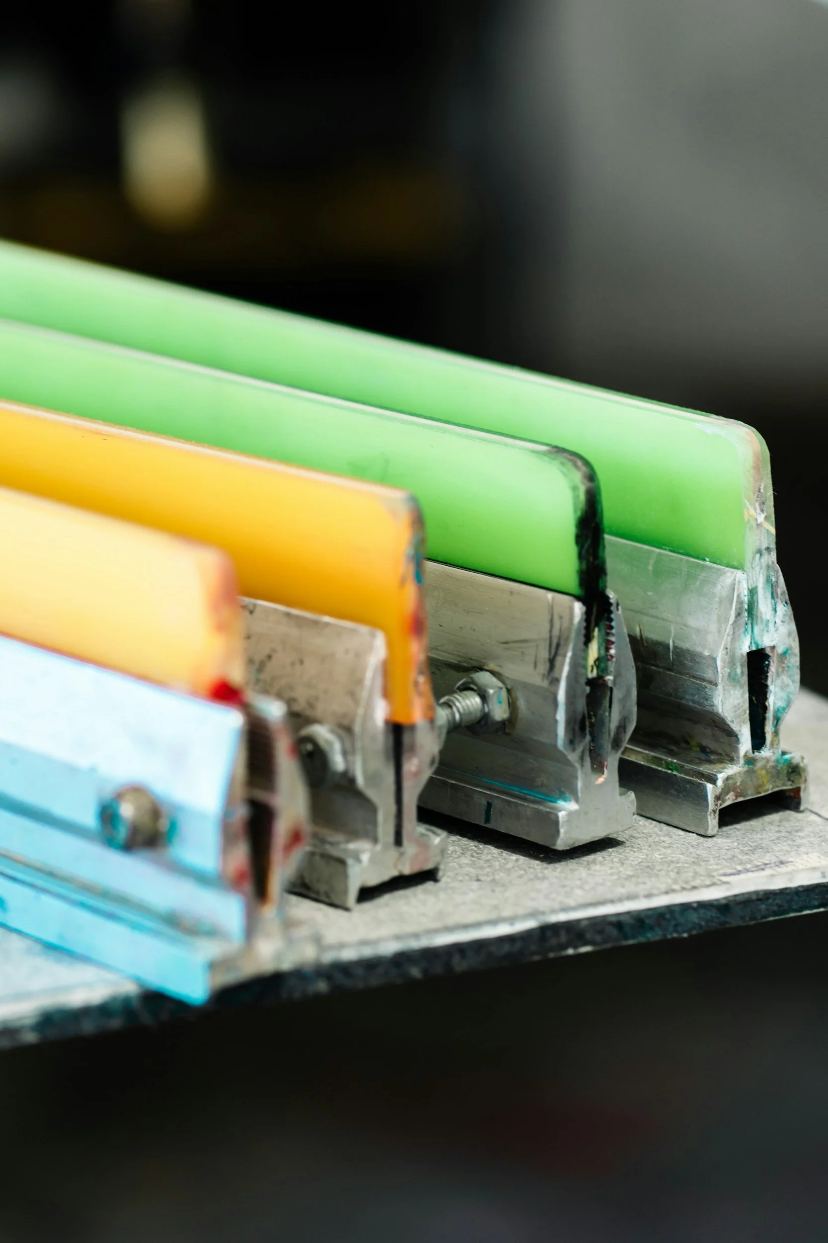 Close-up of screen printing squeegees, with metal bases and visible screws, sitting with some plastisol ink leftover on them.