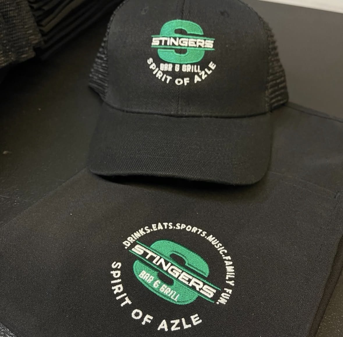 Black baseball cap with a logo that says "Stingers" in white with a green background and the words "Bar & Grill" underneath, with the phrase "Spirit of Azle" below. The cap is placed on a black apron with the logo also embroidered.