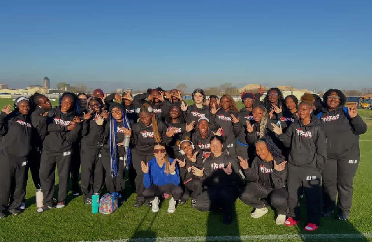 A group of women and girls wearing matching black athletics jackets and pants, standing on a grassy field on a sunny day, posing for a team photo.