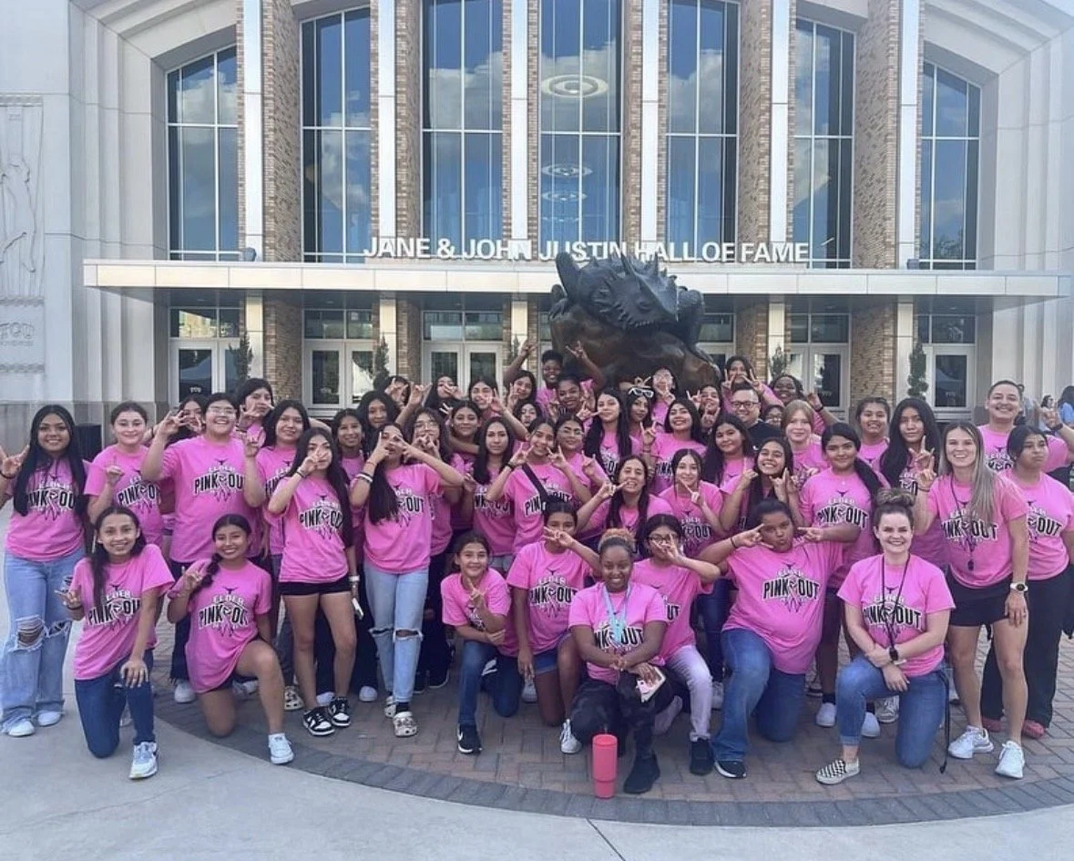 Group of young girls wearing pink shirts for volleyball pink out gathering outside of a building with large glass windows, with TCU stadium in the background.