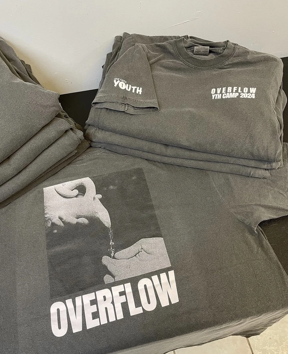 Stack of gray t-shirts, some folded, with a printed t-shirt on top that has "Overflow Youth Camp 2024" written on it, and a gray shirt with a black and white halftone print close-up and the word "Overflow" at the bottom.