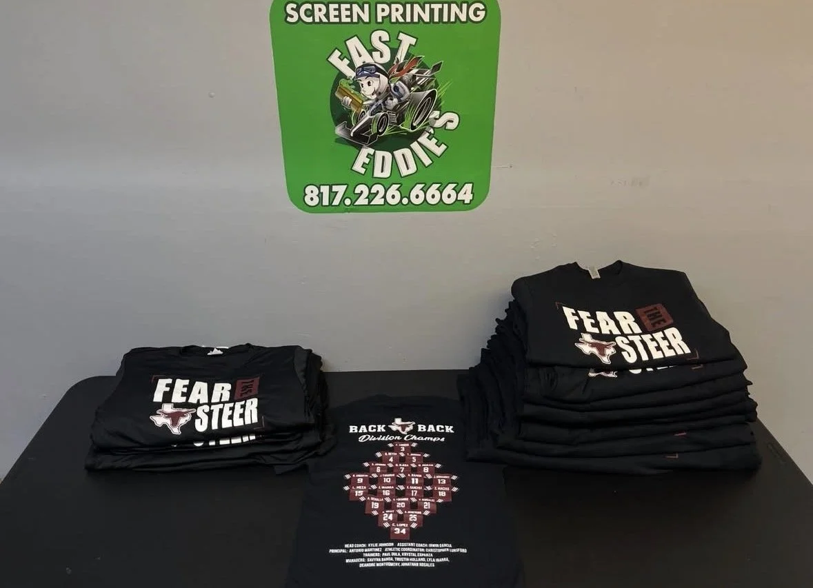 Stacks of black t-shirts with 'FEAR THE STEER' logo and design, displayed on a table with a green sign for 'FAST EDDIES' screen printing and their contact number.
