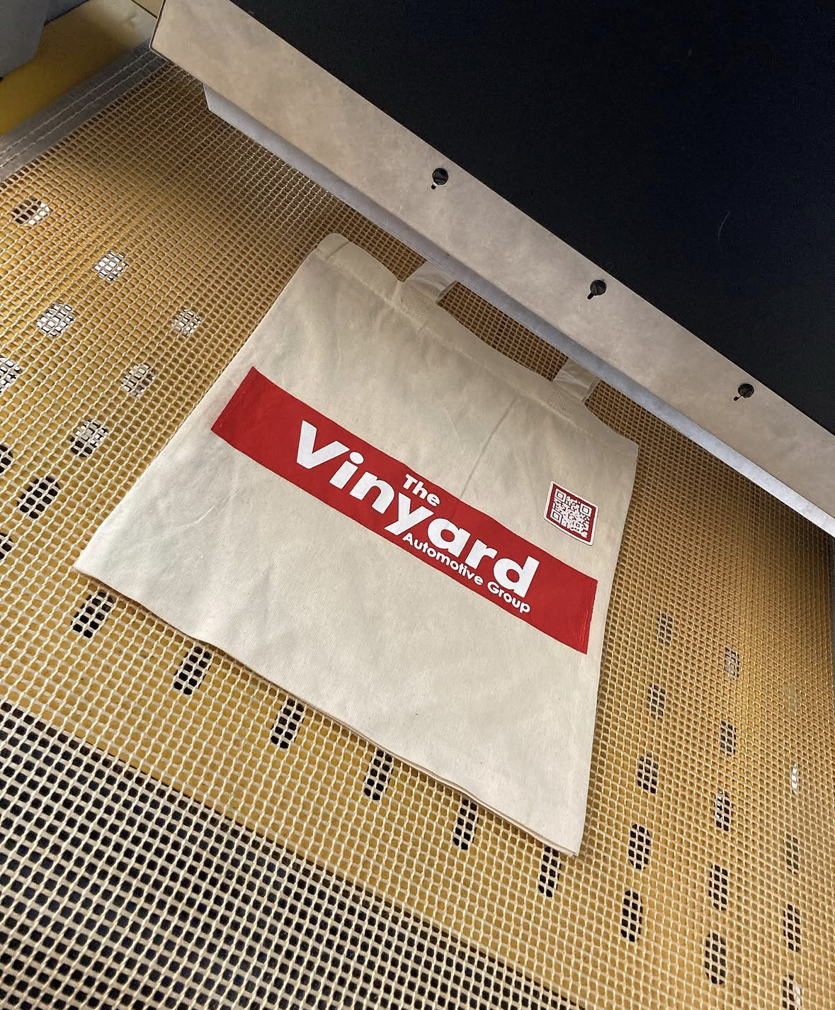 A white tote bag with the red and white logo of The VinYard Automotive Group on a screen printing conveyor dryer
