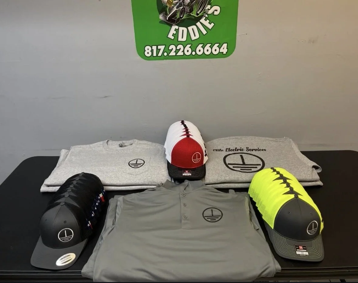 Display of branded clothing and caps with logo, including gray shirts, gray t-shirts, black, white, and neon yellow caps, on a black table with a piece of gray cloth behind, with a green sign above that includes a phone number.