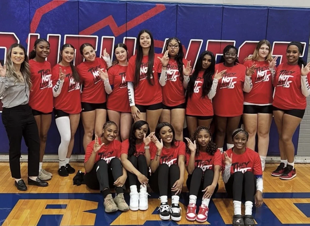 Group of female volleyball players and coach posing on a gymnasium court in front of a backdrop with bold text, dressed in red and black uniforms with some wearing long sleeve shirts. They are making victory signs with their hands, smiling and showing team spirit.