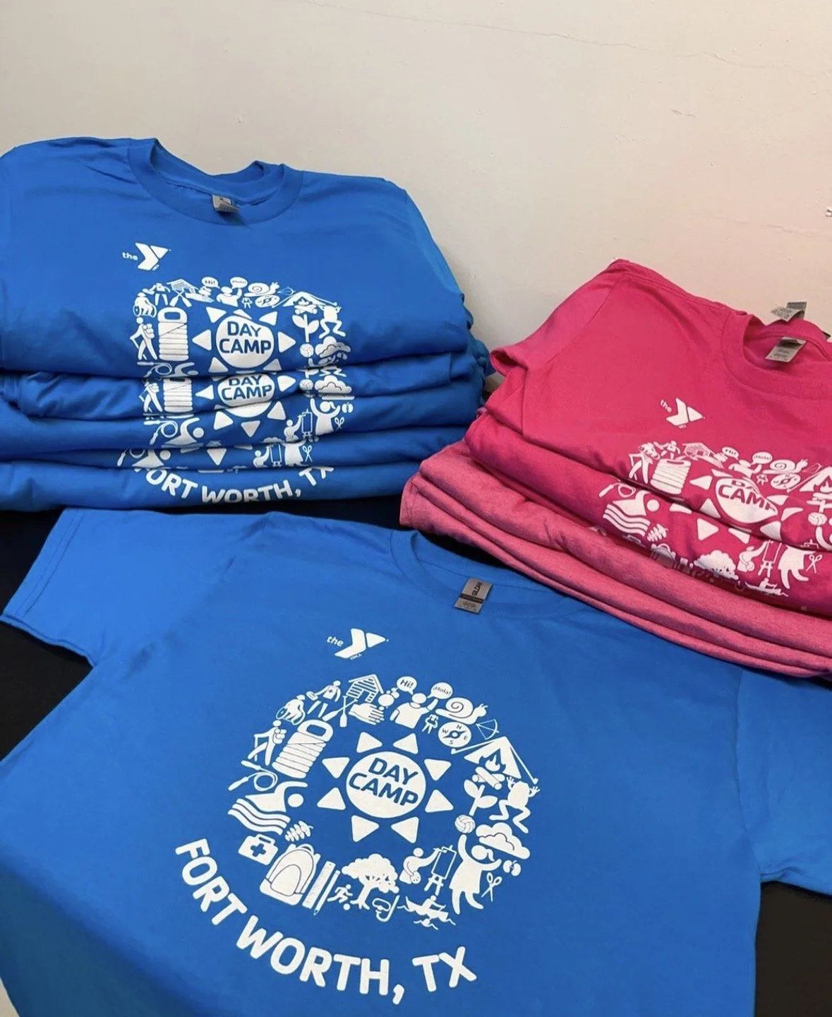 Stacks of blue and pink t-shirts with a white graphic design and text that reads 'YMCA Day Camp Fort Worth, TX' and features icons related to camp activities.