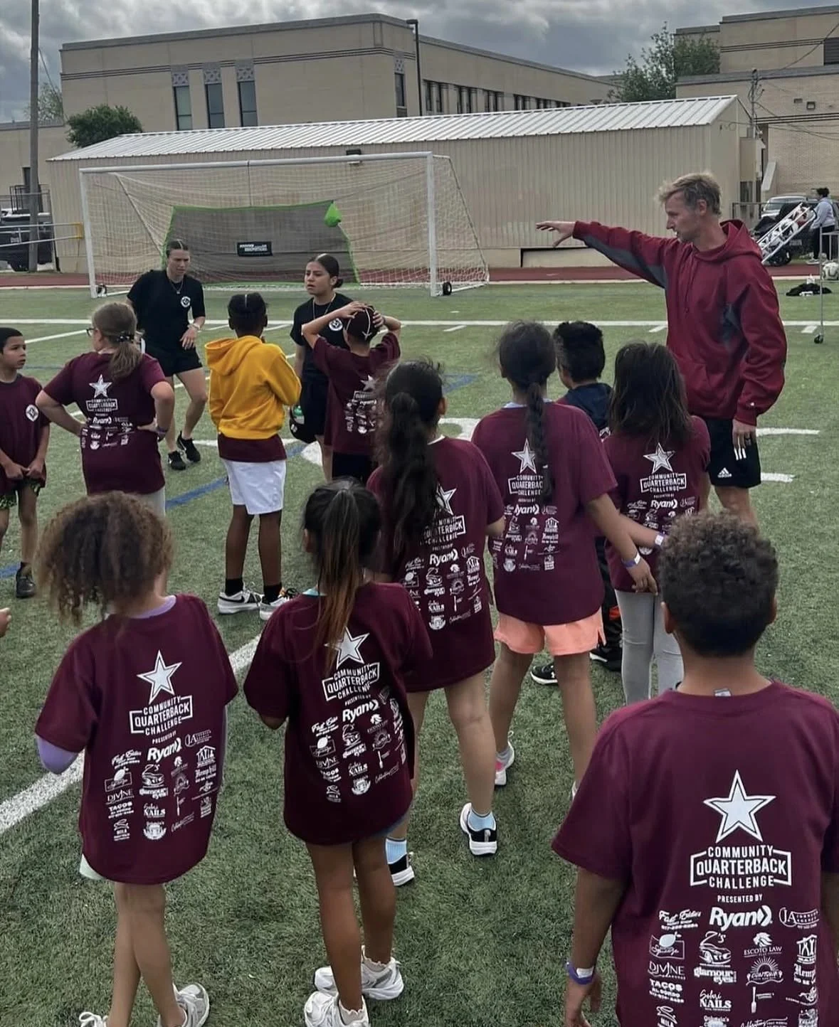 A group of young kids participating in a sports training session on a football field. An adult coach is giving instructions, and a soccer goal is visible in the background. The girls are wearing maroon t-shirts with logos and text on the back.